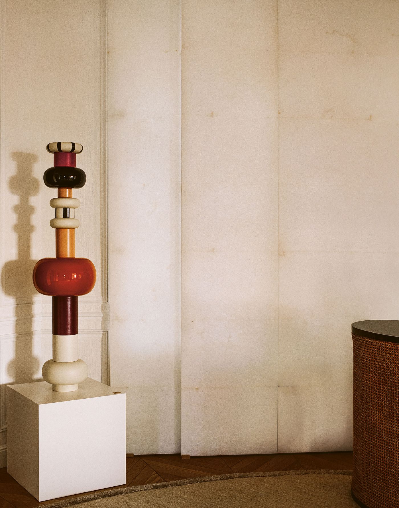 An artistic, stacked sculptural piece in red, black, and cream tones stands prominently on a white plinth, adding a playful yet sophisticated focal point to the room. The background features light, textured wall panels, hinting at a hidden door or feature, and a glimpse of a woven furnishing.