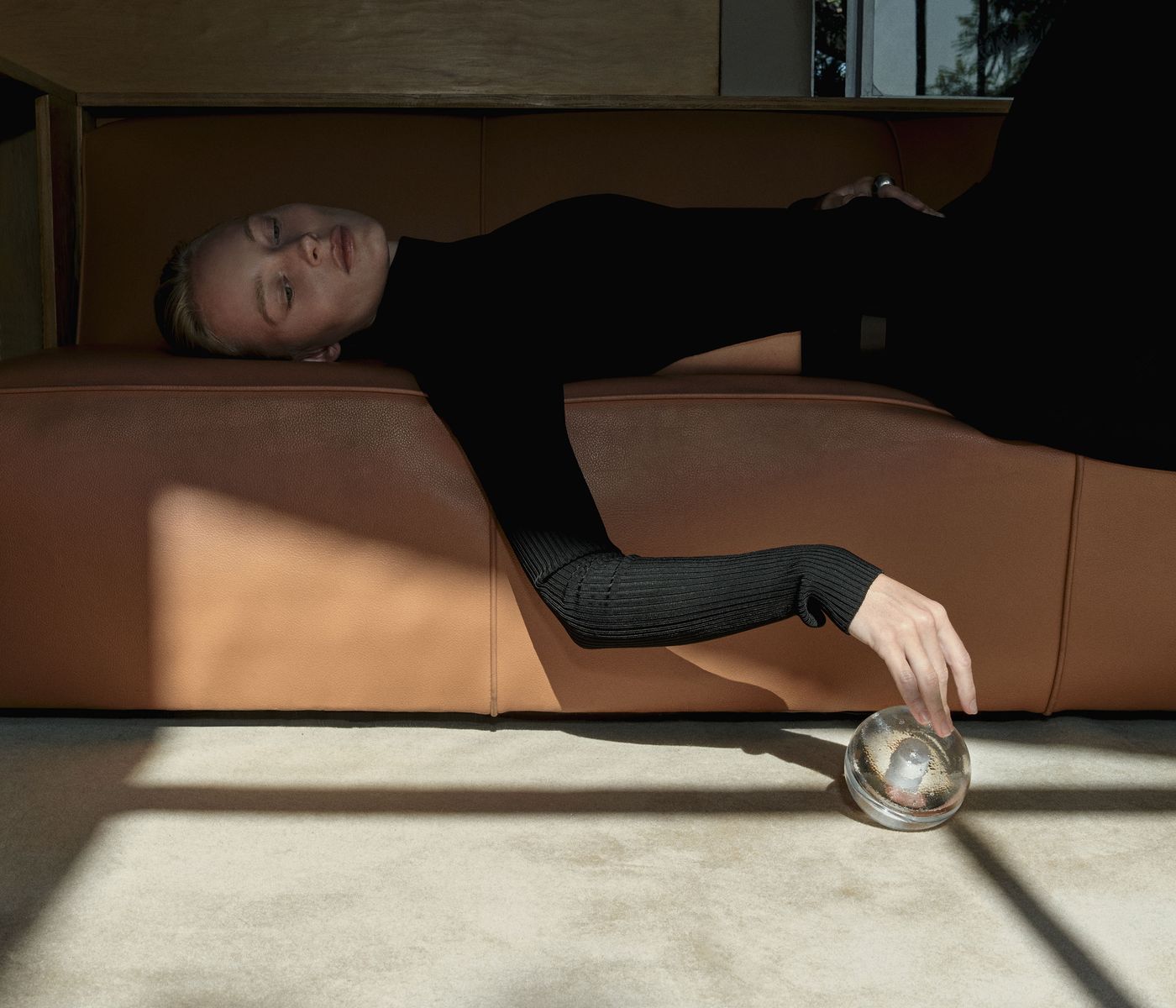 A person dressed in black reclines on a tan leather sofa, extending one arm downward to gently touch a clear, spherical glass object resting on the floor. Sunlight streams in, casting geometric shadows across the scene. The atmosphere is calm and minimalistic