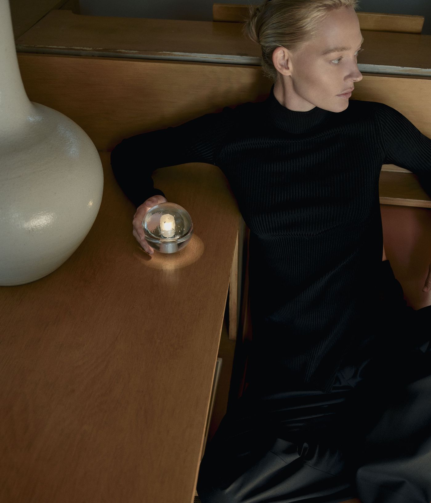 A person in a ribbed black turtleneck sits against a wooden surface, holding a 14p bocci sphere. The setting features warm wood tones and a large white ceramic vase, evoking a serene, intimate mood