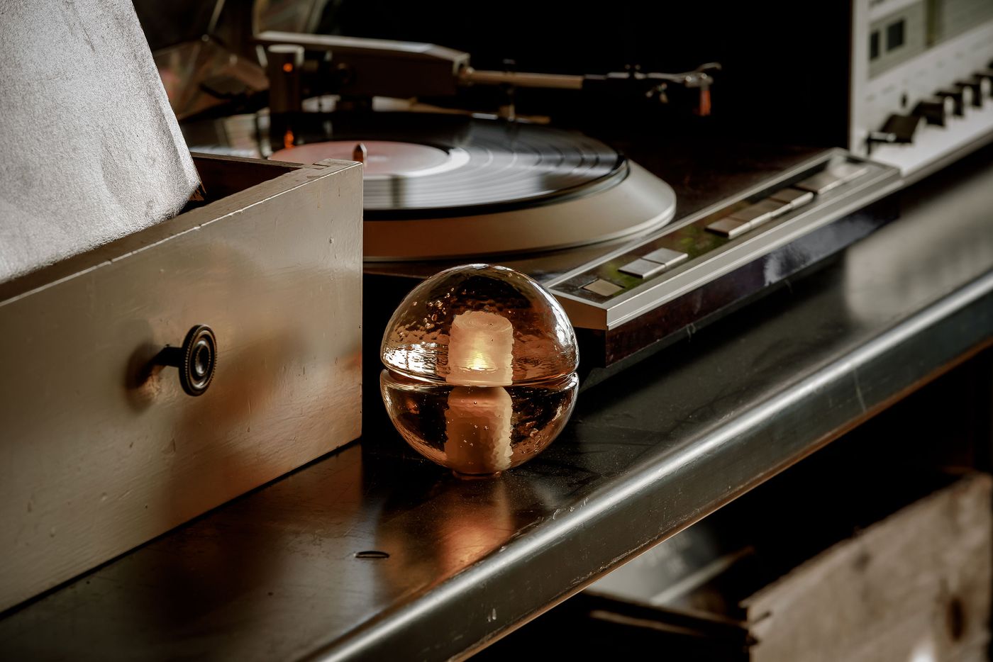 A 14p sphere with small bubbles inside sits on a dark surface next to a vintage record player and wooden drawer. The scene is softly lit, highlighting the orb’s reflective qualities and the retro audio equipment in the background