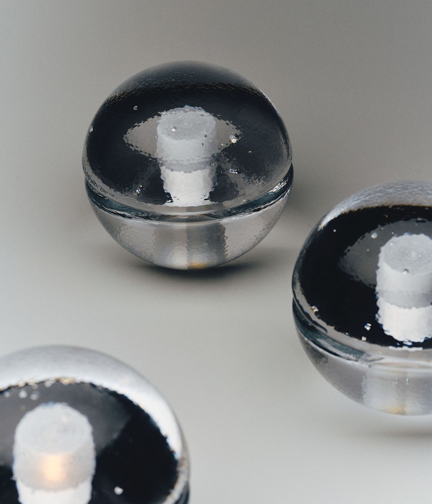 A close-up of several clear, textured glass spheres with visible bubbles, each containing a white candle at its center, arranged on a neutral background. The craftsmanship of the glass is highlighted by the lighting.