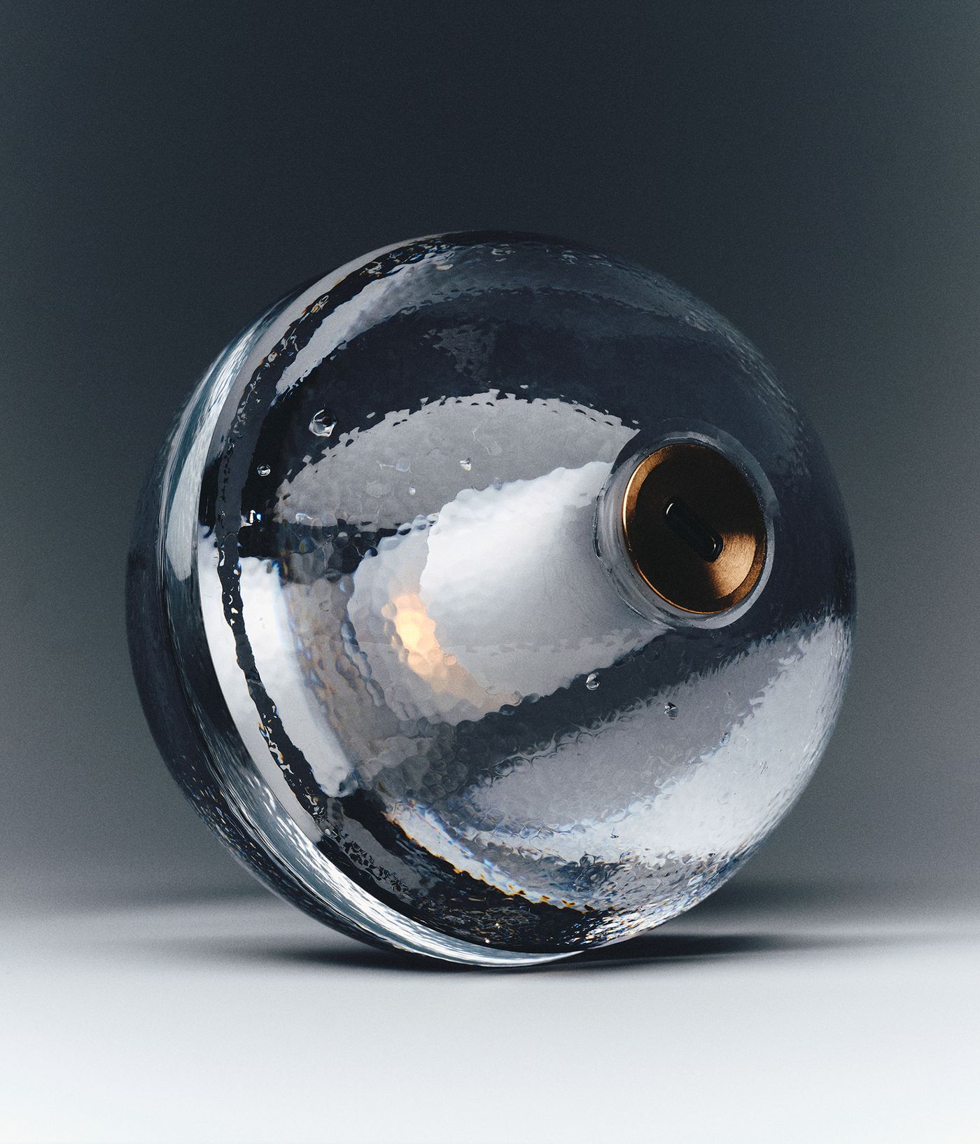 A single, clear, textured 14 p sphere by bocci is shown in close-up against a gradient background. The sphere is slightly tilted, revealing its metallic base and the subtle play of light and shadow on its surface