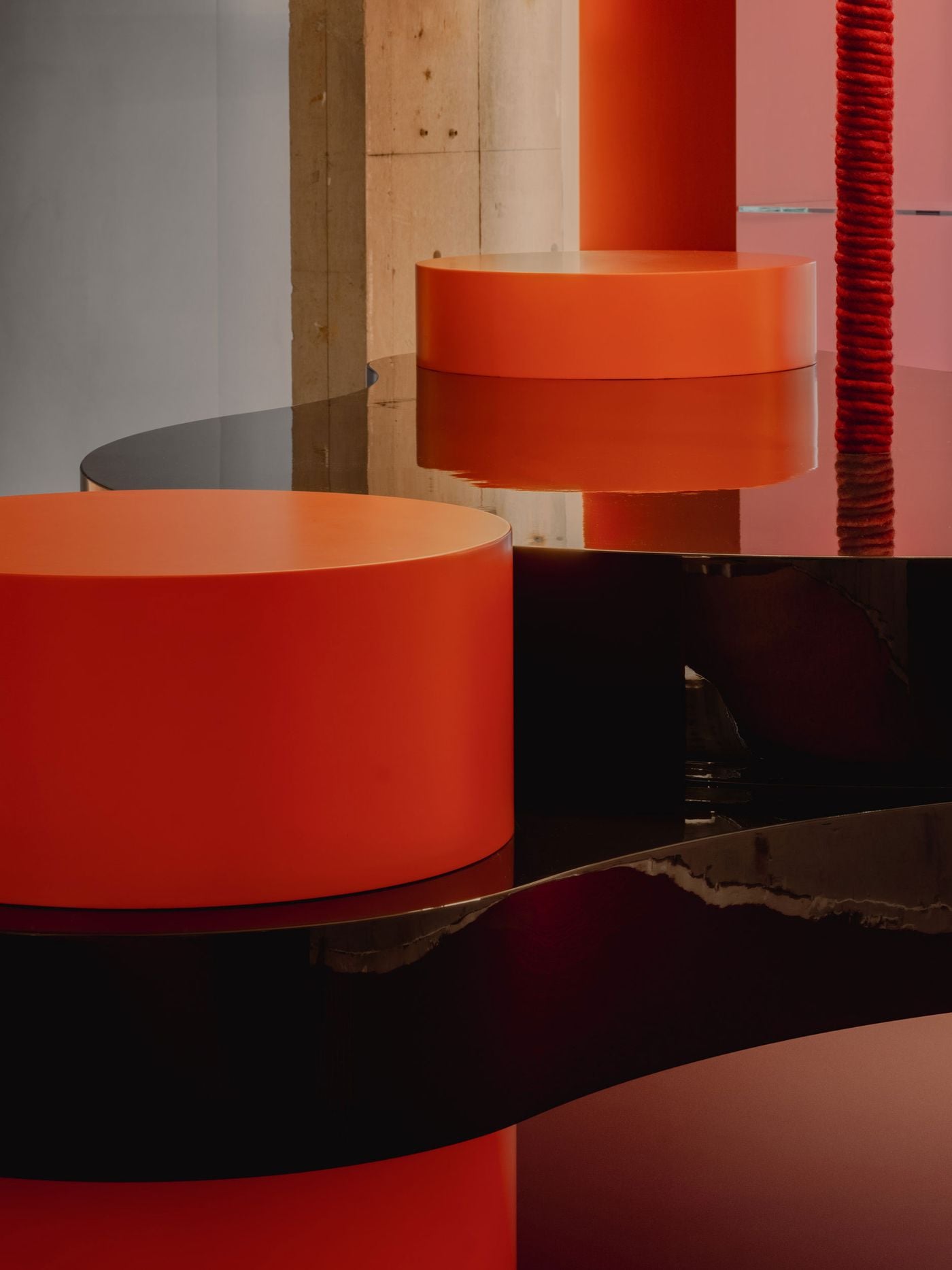 Glossy black surfaces loop around cylindrical orange plinths in a close-up of the ground-floor display counter. Reflections distort the forms and intensify the saturated red-orange palette, while a raw concrete column and rope-wrapped vertical element underscore I IN’s recurring contrast between polished intervention and unfinished structure.
