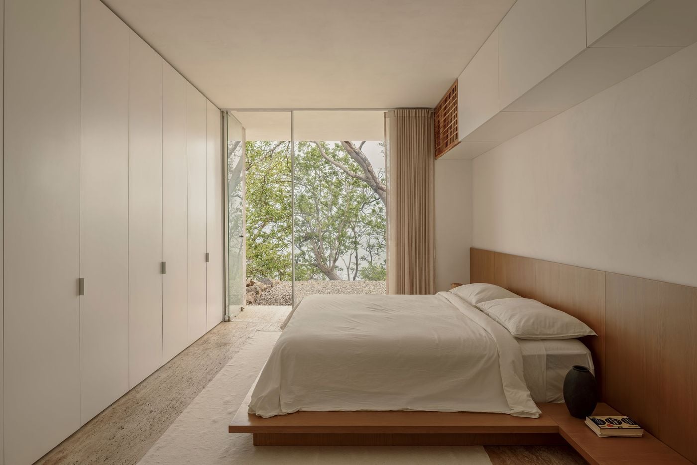A low timber platform bed with an extended headboard panel and integrated bedside shelf anchors this bedroom, opposite a wall of flush white wardrobes. A slatted timber grille and linen curtain soften the room, while full-height glazing opens onto a rocky garden beneath a leafy canopy. A low timber platform bed with an extended headboard panel and integrated bedside shelf anchors this bedroom, opposite a wall of flush white wardrobes. A slatted timber grille and linen curtain soften the room, while full-height glazing opens onto a rocky garden beneath a leafy canopy.