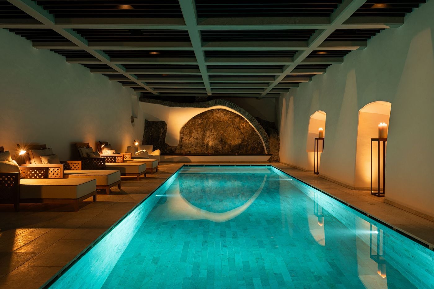 The indoor pool at Castel Badia's spa runs beneath a sage-painted coffered timber ceiling towards a vaulted niche of exposed bedrock. Green stone tiles turn the water a vivid turquoise, while lattice-framed loungers line one side and candle lanterns glow within arched recesses opposite. The indoor pool at Castel Badia's spa runs beneath a sage-painted coffered timber ceiling towards a vaulted niche of exposed bedrock. Green stone tiles turn the water a vivid turquoise, while lattice-framed loungers line one side and candle lanterns glow within arched recesses opposite.