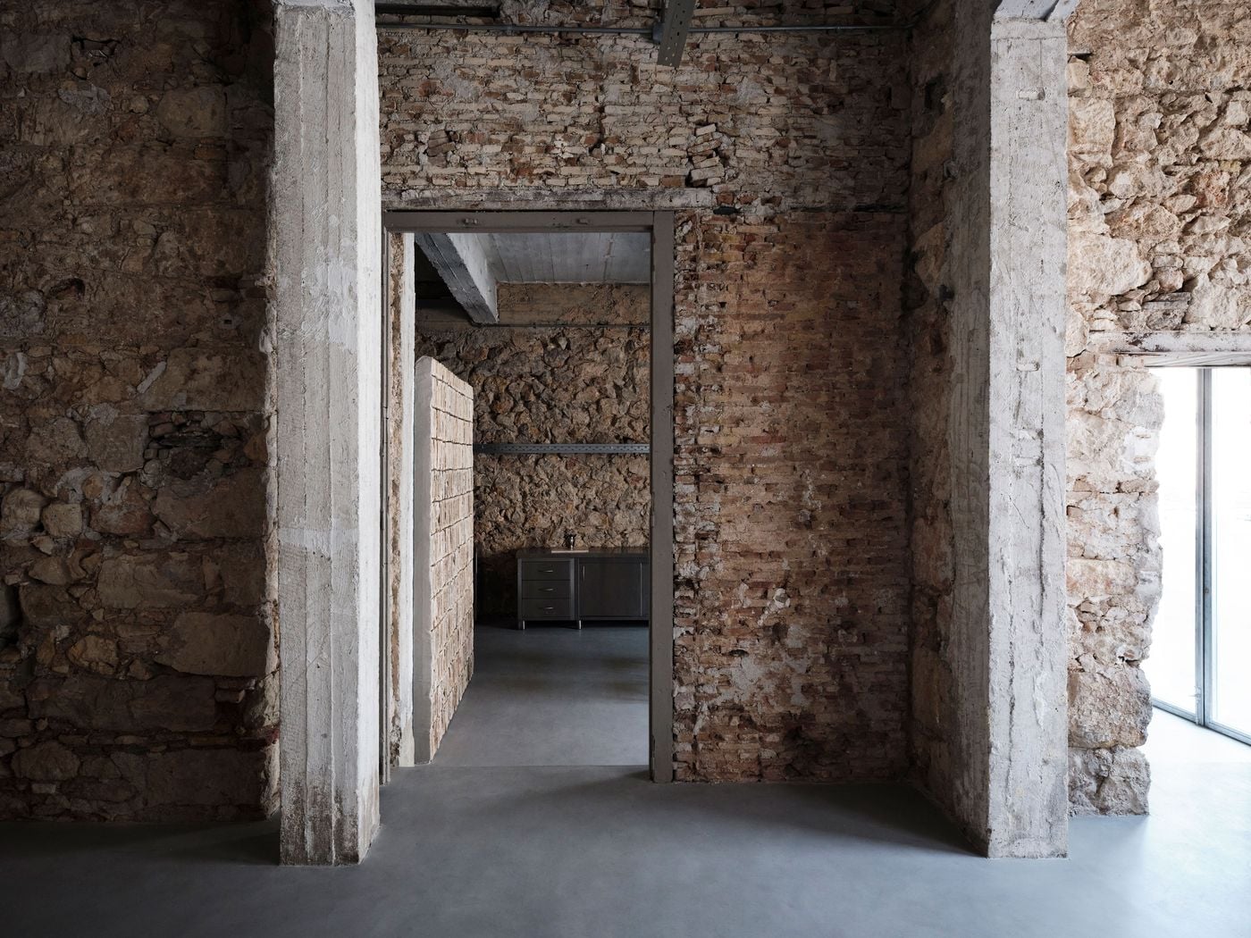 Exposed stone, brick and concrete columns frame a sequence of rooms connected by reopened original doorways. The uninterrupted grey floor lends visual coherence to the varied masonry, while daylight entering from the right accentuates the irregular surfaces and evidence of the warehouse’s successive alterations. Exposed stone, brick and concrete columns frame a sequence of rooms connected by reopened original doorways. The uninterrupted grey floor lends visual coherence to the varied masonry, while daylight entering from the right accentuates the irregular surfaces and evidence of the warehouse’s successive alterations.