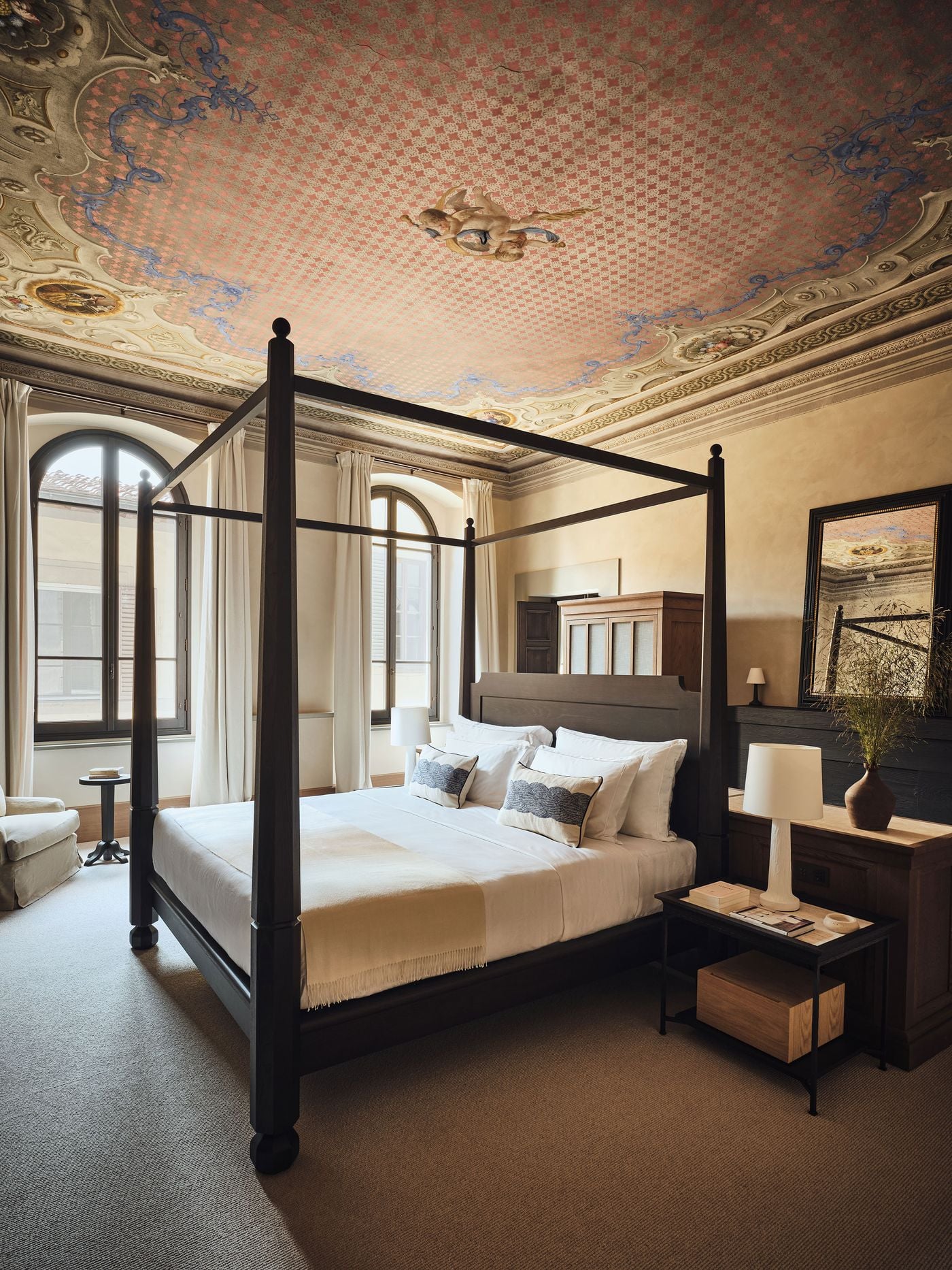 A painted ceiling of rose lattice, blue ribbon scrolls, rocaille cartouches and floating putti crowns a bedroom at La Réserve Firenze. Below, a dark four-poster bed with ball finials, crisp white linen and plaster-textured lamps keeps the room grounded, while arched windows draw in the light from the street. A painted ceiling of rose lattice, blue ribbon scrolls, rocaille cartouches and floating putti crowns a bedroom at La Réserve Firenze. Below, a dark four-poster bed with ball finials, crisp white linen and plaster-textured lamps keeps the room grounded, while arched windows draw in the light from the street.