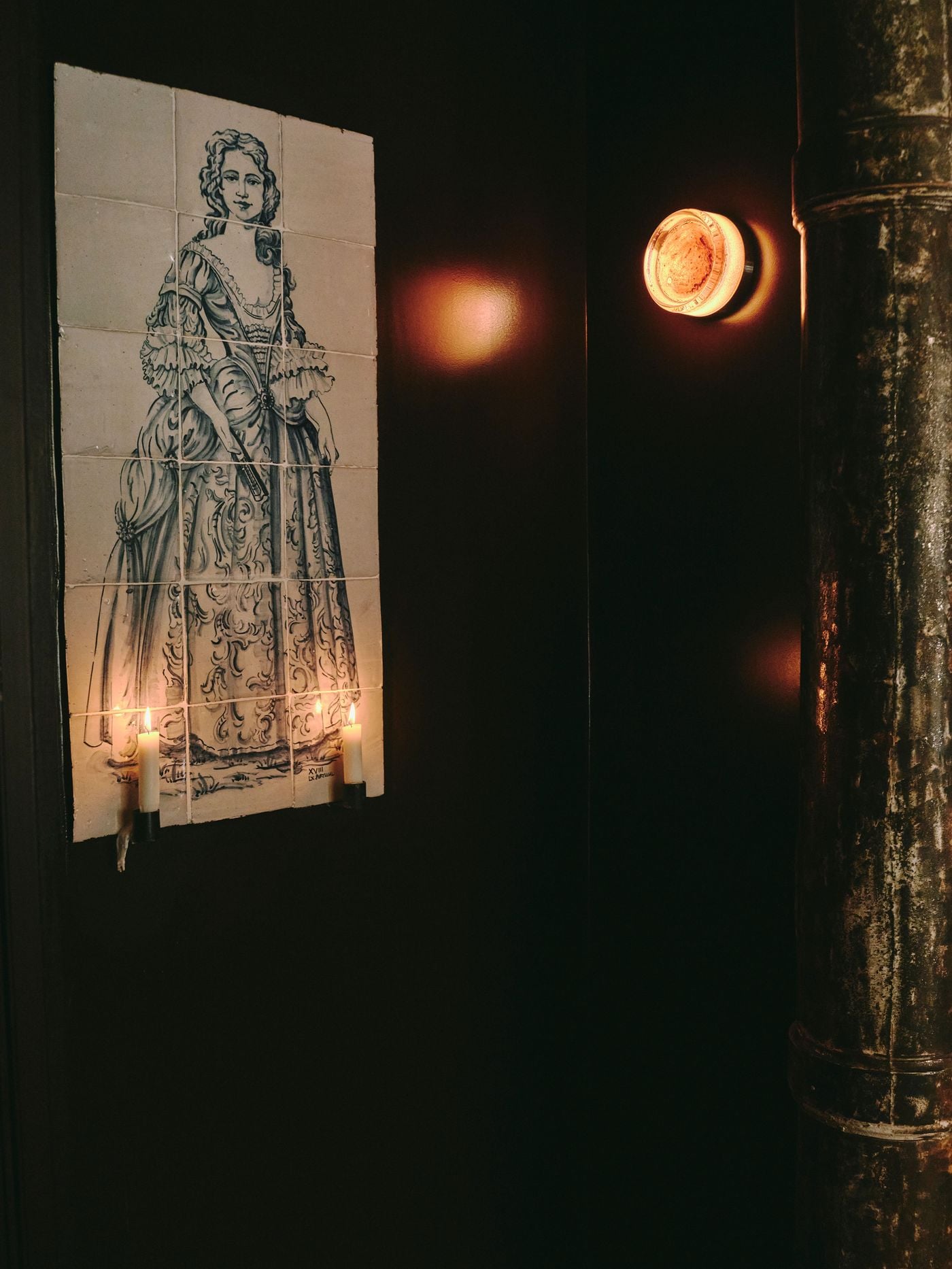 A tiled portrait of an 18th-century lady in a ruffled gown emerges from the darkness at SaltoBar in Paris, lit by two candles fixed to its base. Nearby, a thick glass wall light casts an amber halo onto deep brown walls. Beside it rises a cast-iron column, its paint worn to a mottled patina. A tiled portrait of an 18th-century lady in a ruffled gown emerges from the darkness at SaltoBar in Paris, lit by two candles fixed to its base. Nearby, a thick glass wall light casts an amber halo onto deep brown walls. Beside it rises a cast-iron column, its paint worn to a mottled patina.