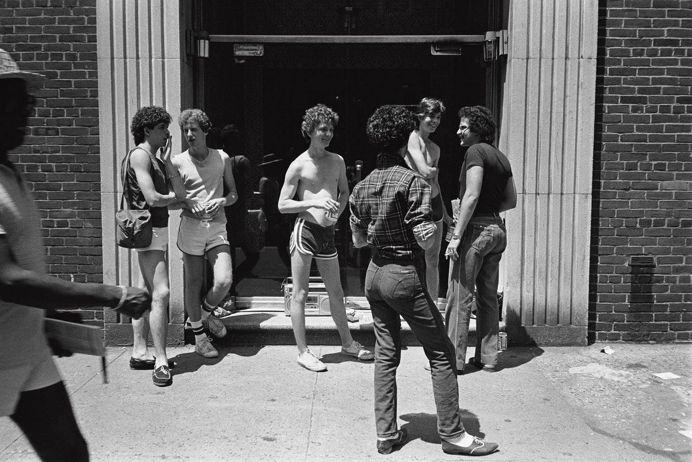 A group of young men loiters in the doorway of a brick building framed by fluted stone pilasters, dressed in short running shorts, tank tops, plaid flannel and high-waisted jeans, a boombox at their feet. A blurred passer-by strides through the foreground of this sunlit street scene from Don't Worry Boys. A group of young men loiters in the doorway of a brick building framed by fluted stone pilasters, dressed in short running shorts, tank tops, plaid flannel and high-waisted jeans, a boombox at their feet. A blurred passer-by strides through the foreground of this sunlit street scene from Don't Worry Boys.