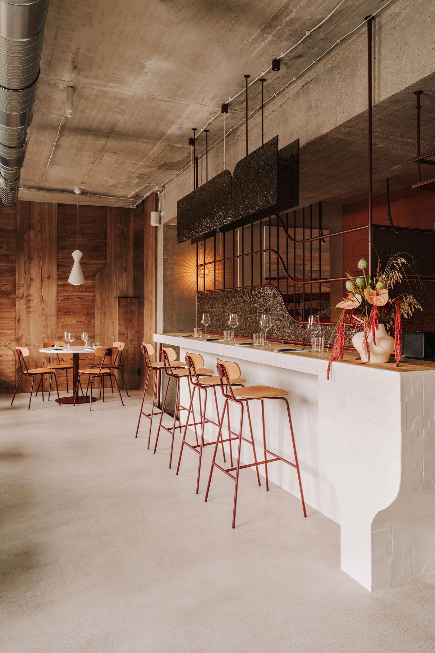 A wider view of WIN’s upstairs dining area reveals the sculptural white-tiled counter with its curved recesses, accompanied by bentwood stools on burgundy metal frames. Sinuous metalwork, textured glass partitions and a suspended dark canopy contrast with raw concrete surfaces, while timber-clad walls and a small dining table introduce warmth to the industrial interior. A wider view of WIN’s upstairs dining area reveals the sculptural white-tiled counter with its curved recesses, accompanied by bentwood stools on burgundy metal frames. Sinuous metalwork, textured glass partitions and a suspended dark canopy contrast with raw concrete surfaces, while timber-clad walls and a small dining table introduce warmth to the industrial interior.