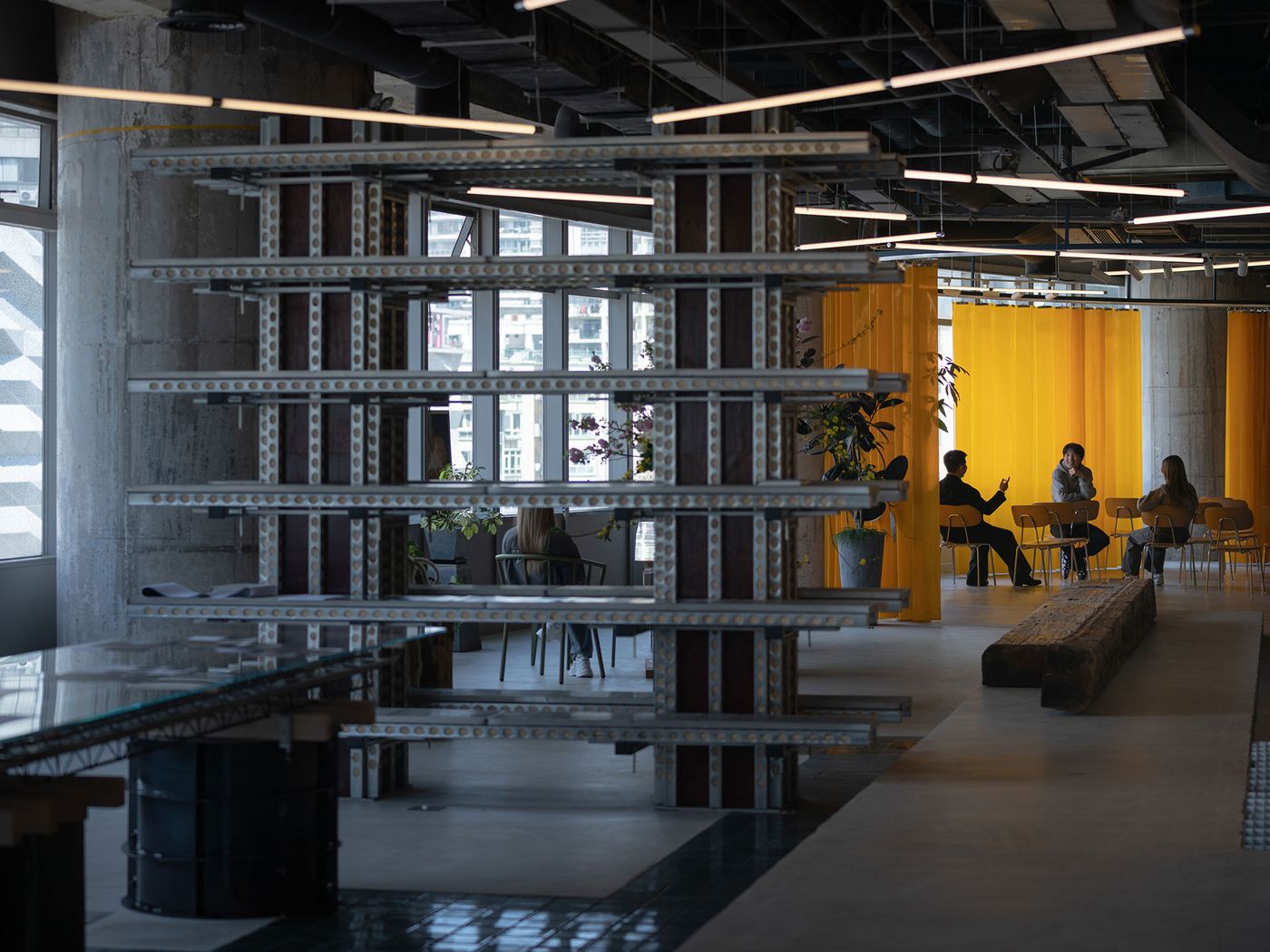 Natural light illuminates the versatile 889 GLO Art Space, highlighting the industrial shelving unit and a rustic wooden bench. Beyond, a warm yellow curtain encloses a seating area where people are gathered, illustrating the harmonious blend of raw materials and vibrant color, defining distinct zones within the spacious, multi-functional design.