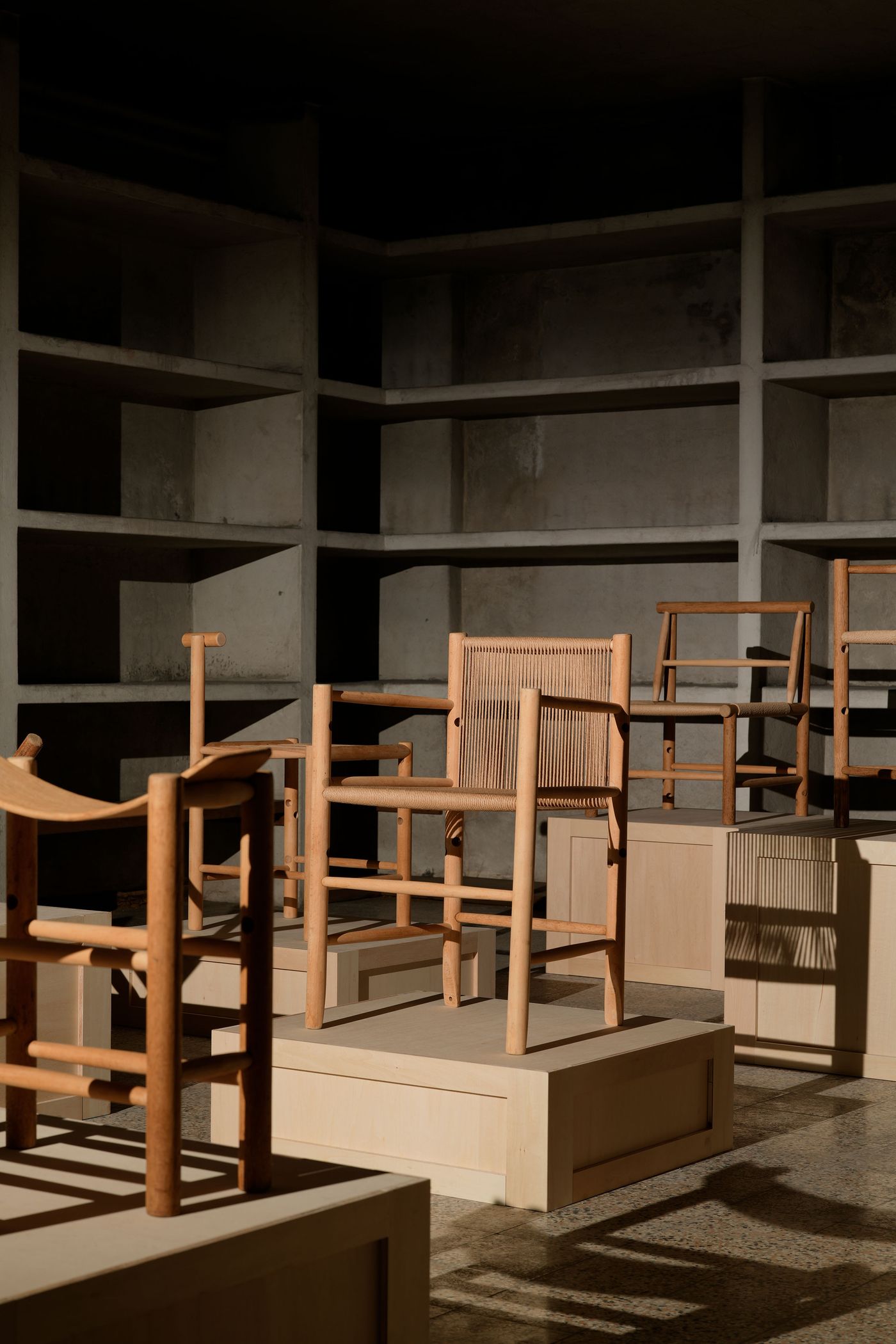 A minimalist display of wooden chairs on elevated platforms, set against a concrete backdrop with soft lighting and shadows.
