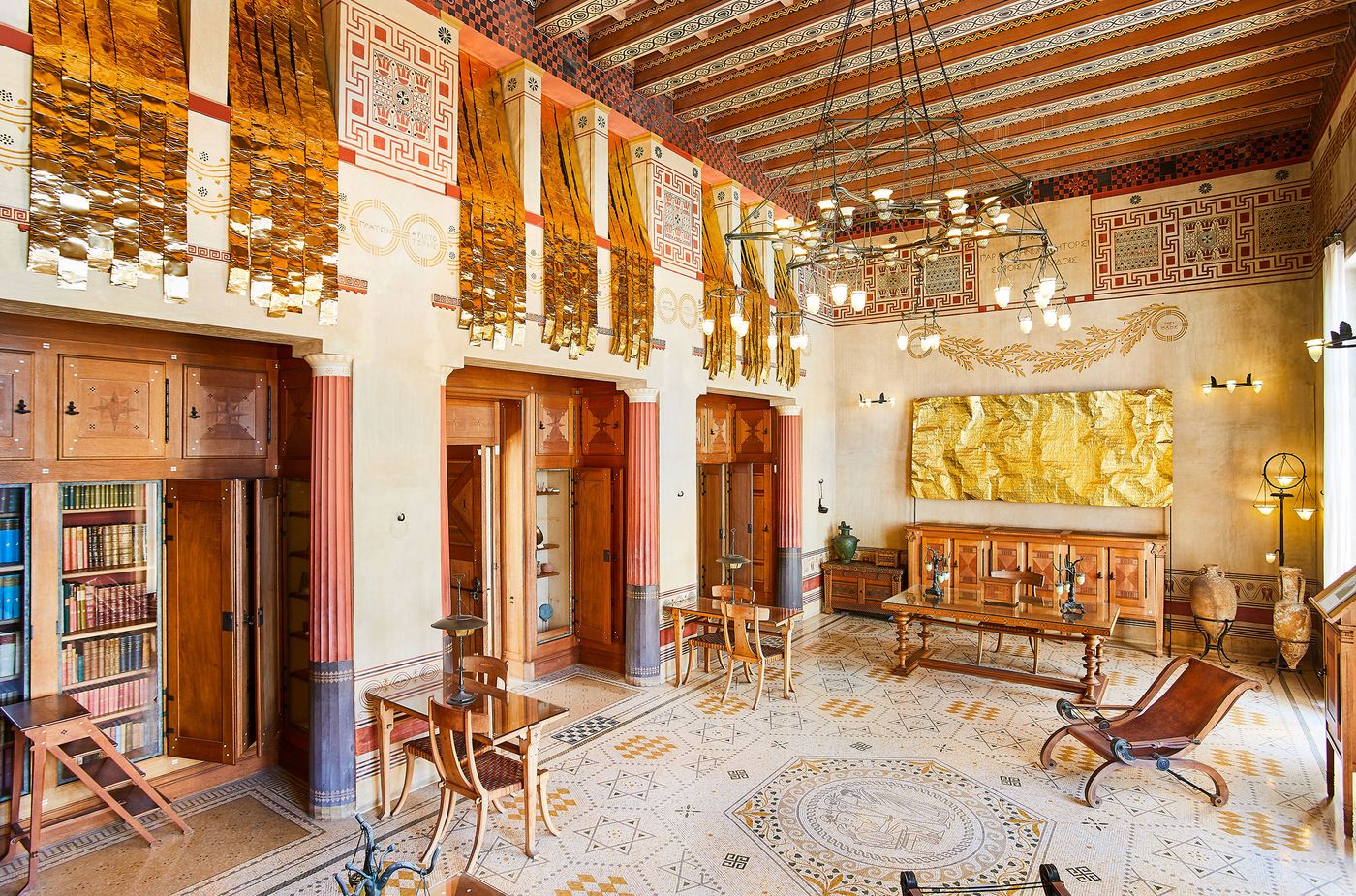 An expansive view of an interior space at Villa Kerylos. The room features a complex mosaic floor, a coffered wood ceiling, and walls with intricate murals. Two modern art pieces by Gabriel Leger—a large, crinkled gold sheet and a suspended, shimmering installation—are placed alongside traditional furniture and a bookshelf. An expansive view of an interior space at Villa Kerylos. The room features a complex mosaic floor, a coffered wood ceiling, and walls with intricate murals. Two modern art pieces by Gabriel Leger—a large, crinkled gold sheet and a suspended, shimmering installation—are placed alongside traditional furniture and a bookshelf.