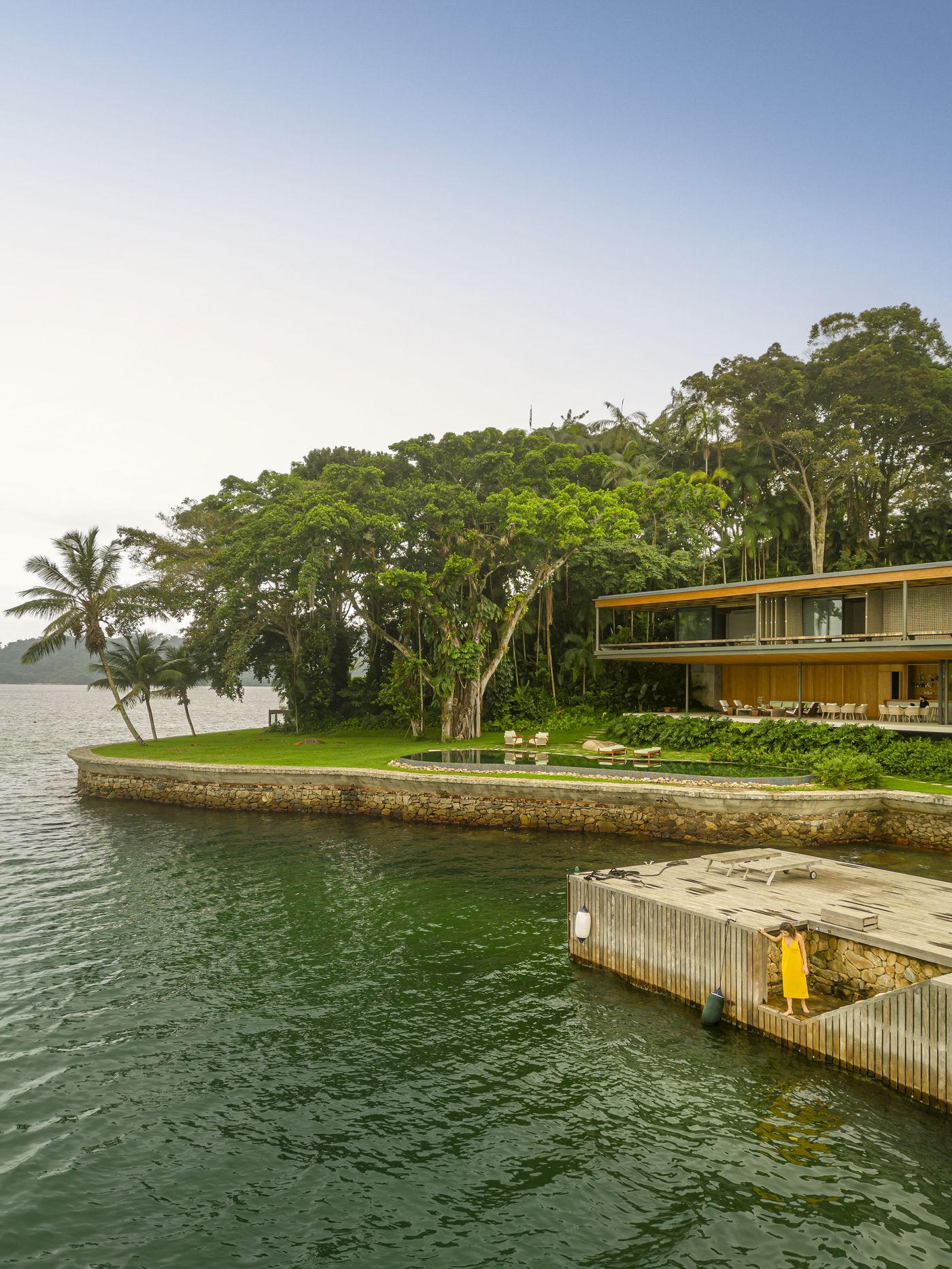 The expansive residence, with its horizontal lines and warm wooden facades, gracefully stretches along the verdant shoreline, overlooking the placid emerald waters. A sleek swimming pool and outdoor lounge area connect seamlessly with the natural landscape, featuring a large, mature tree that anchors the biophilic design to its coastal environment. The expansive residence, with its horizontal lines and warm wooden facades, gracefully stretches along the verdant shoreline, overlooking the placid emerald waters. A sleek swimming pool and outdoor lounge area connect seamlessly with the natural landscape, featuring a large, mature tree that anchors the biophilic design to its coastal environment.