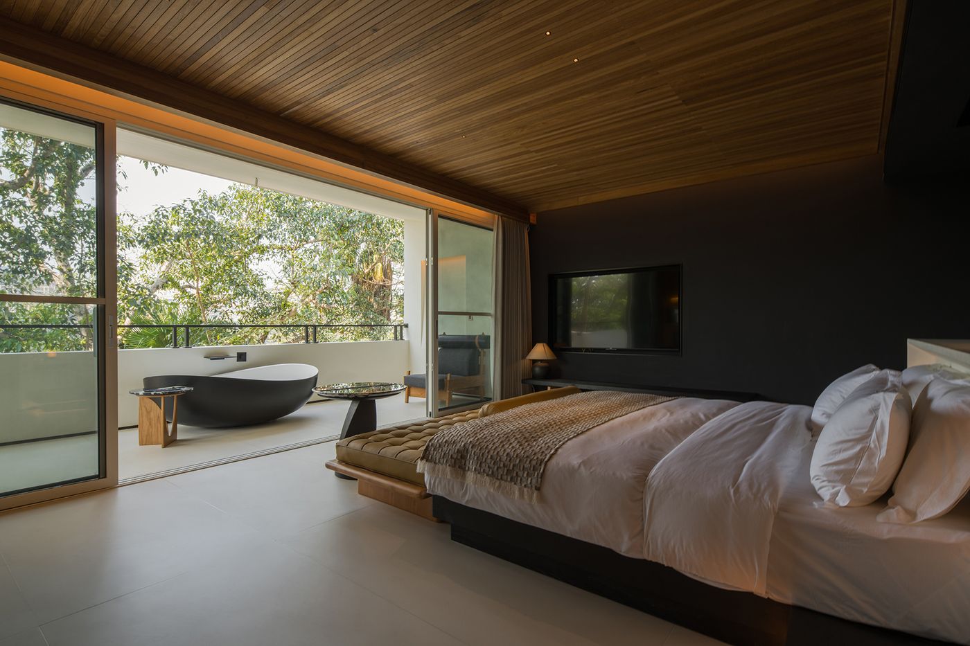 Modern bedroom featuring a large bed, glass walls opening to a balcony with a sleek bathtub and lush greenery outside. Modern bedroom featuring a large bed, glass walls opening to a balcony with a sleek bathtub and lush greenery outside.