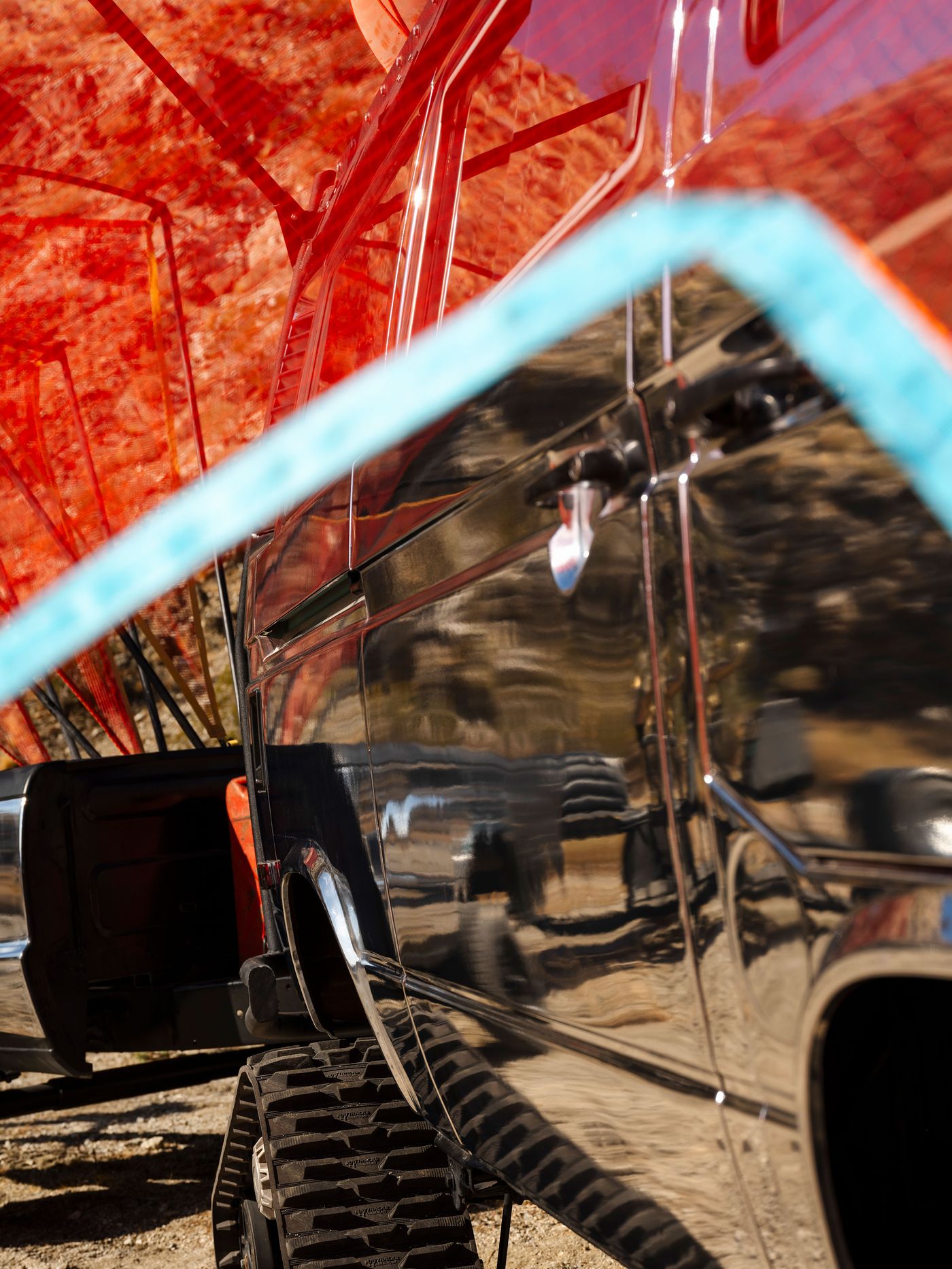 A close-up of a reflective vehicle, showing rugged terrain tires and vibrant red nets overhead, amidst a desert landscape.