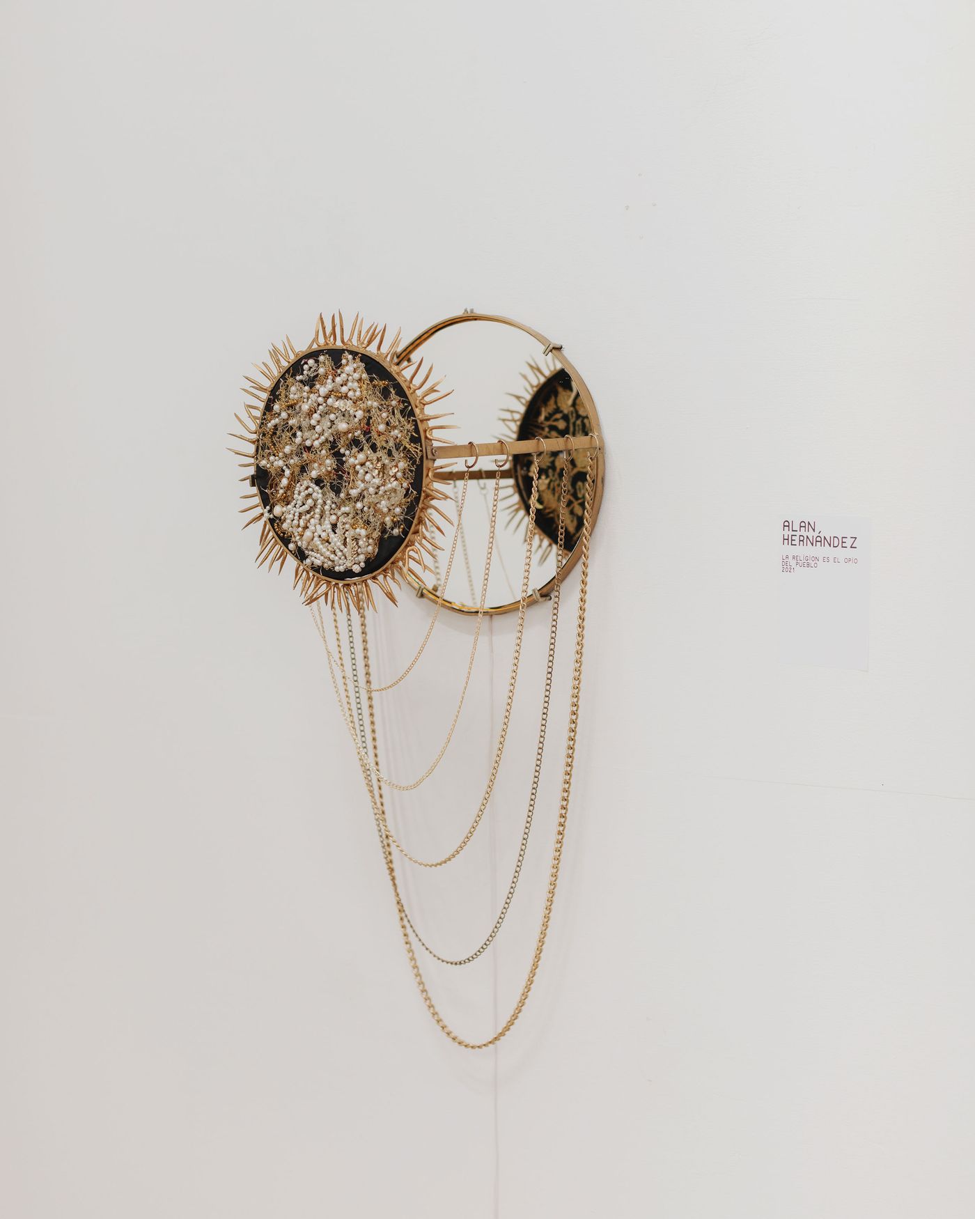 A circular wall art piece by Alan Hernández featuring spikes, beads, chains, and a small mirror, mounted on a white wall.