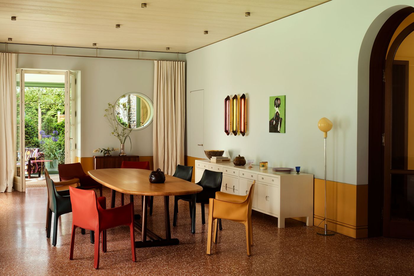 Bright dining room featuring a wooden table, colorful chairs, and a lovely garden view through open doors. Decorative art and plants accentuate the space. Bright dining room featuring a wooden table, colorful chairs, and a lovely garden view through open doors. Decorative art and plants accentuate the space.