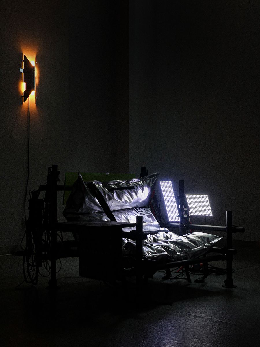 A dimly lit room featuring an industrial-style bed with metallic pillows, flanked by two bright lights, creating a stark contrast. A dimly lit room featuring an industrial-style bed with metallic pillows, flanked by two bright lights, creating a stark contrast.