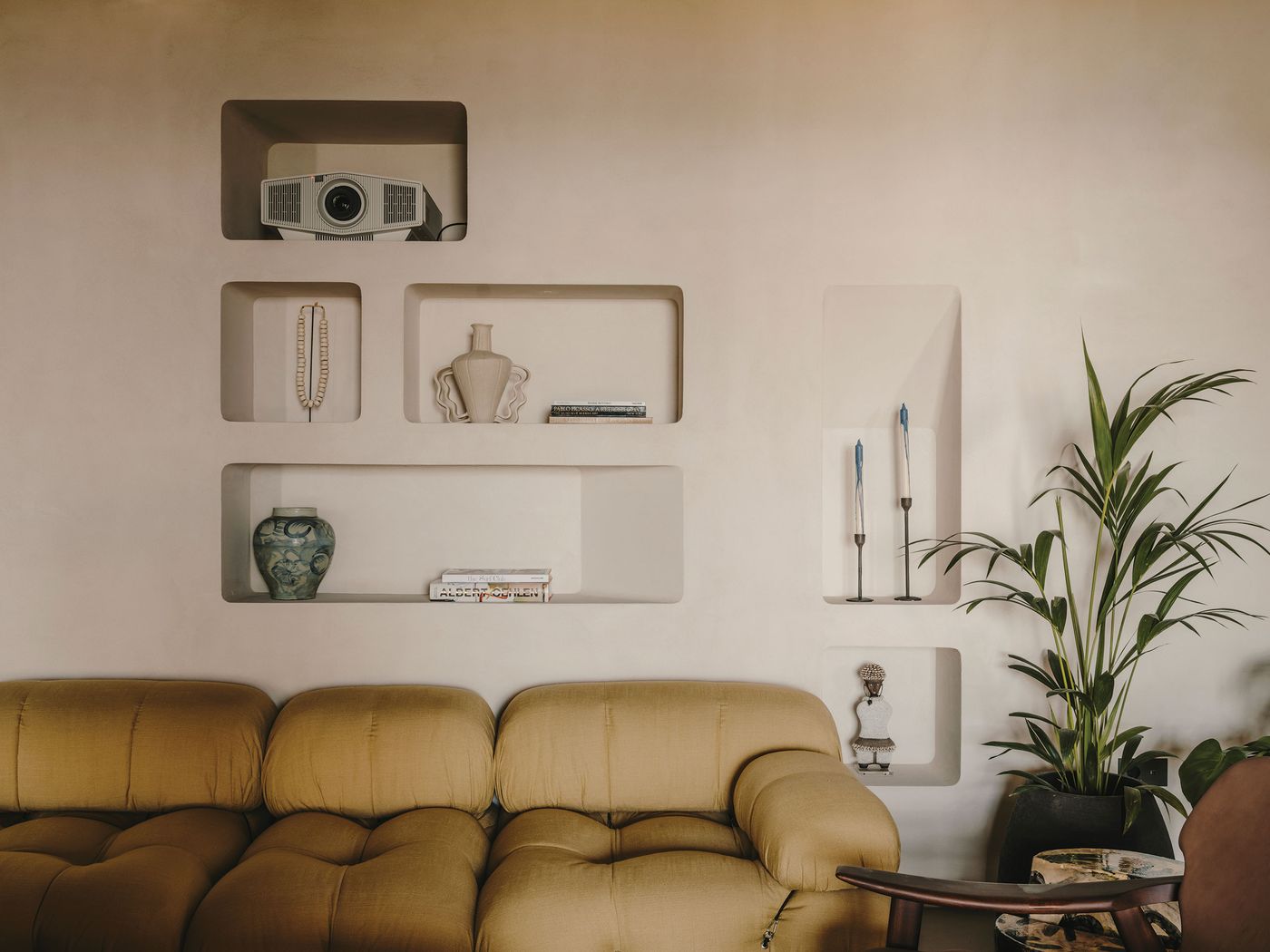 A cozy living room with a mustard yellow sofa, a wall with decorative shelves, and potted plants, featuring a projector and elegant decor items.