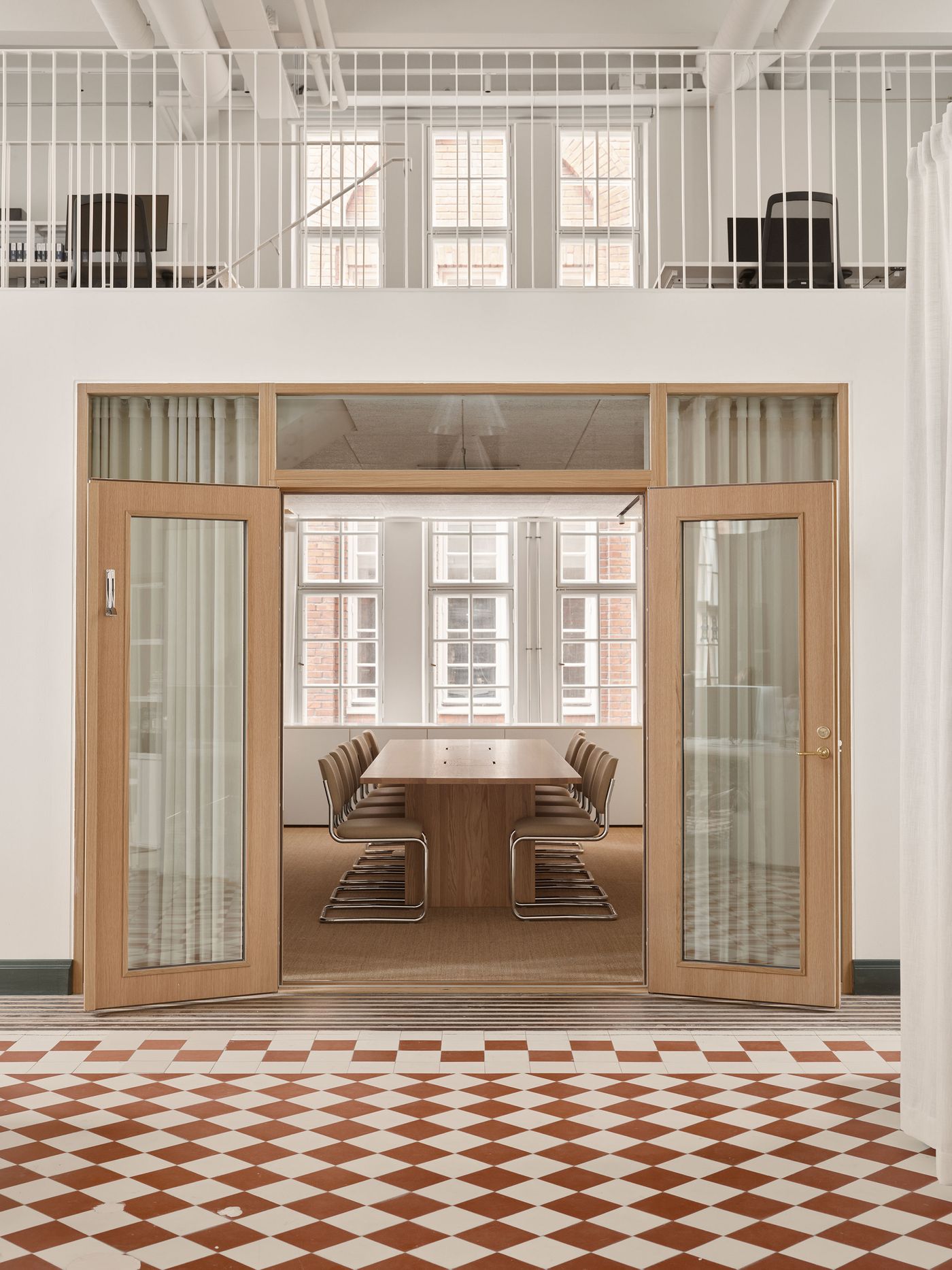 A large meeting room with a long wooden table and modern chairs is framed by a broad wooden archway with glass doors and sheer curtains, hinting at privacy within an open office plan. A mezzanine level with a white railing overlooks the space, blending industrial and refined elements. A large meeting room with a long wooden table and modern chairs is framed by a broad wooden archway with glass doors and sheer curtains, hinting at privacy within an open office plan. A mezzanine level with a white railing overlooks the space, blending industrial and refined elements.
