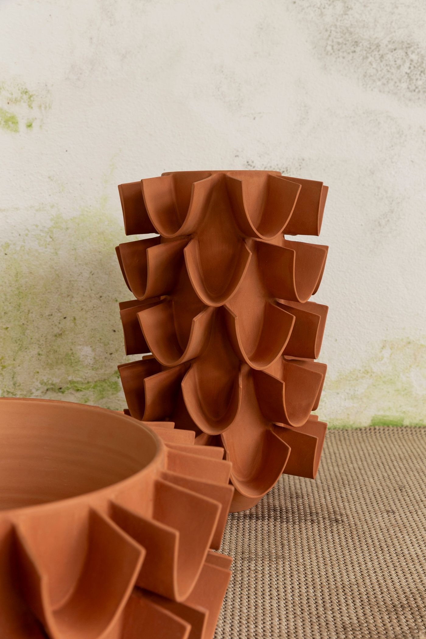 A close-up shot focuses on terracotta ceramic objects with a distinctive scalloped, almost floral design. The main piece is a stack of modular elements forming a tall, textured cylinder. Another similar, but larger, element is partially visible in the foreground. The warm, earthy tone of the clay and the intricate, repeating pattern create a tactile and visually engaging composition against a textured neutral background.