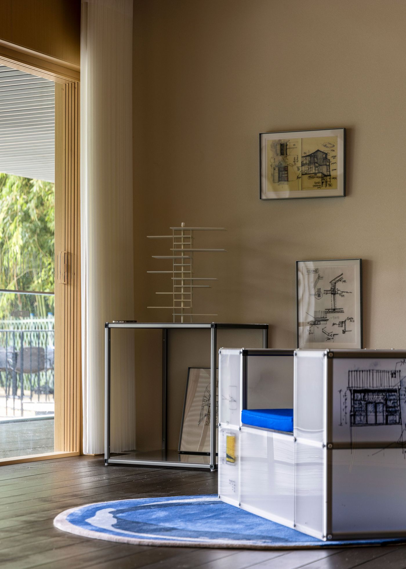 An interior corner of Wutopia Lab's Lake House reveals an eclectic yet refined design. Dark wooden floors ground the space, while a vibrant blue circular rug provides a focal point. Contemporary furniture, including a transparent console table with a minimalist sculpture and a modular white seating unit with a bright blue cushion, complements framed architectural sketches on the wall, adding artistic depth to the room.
