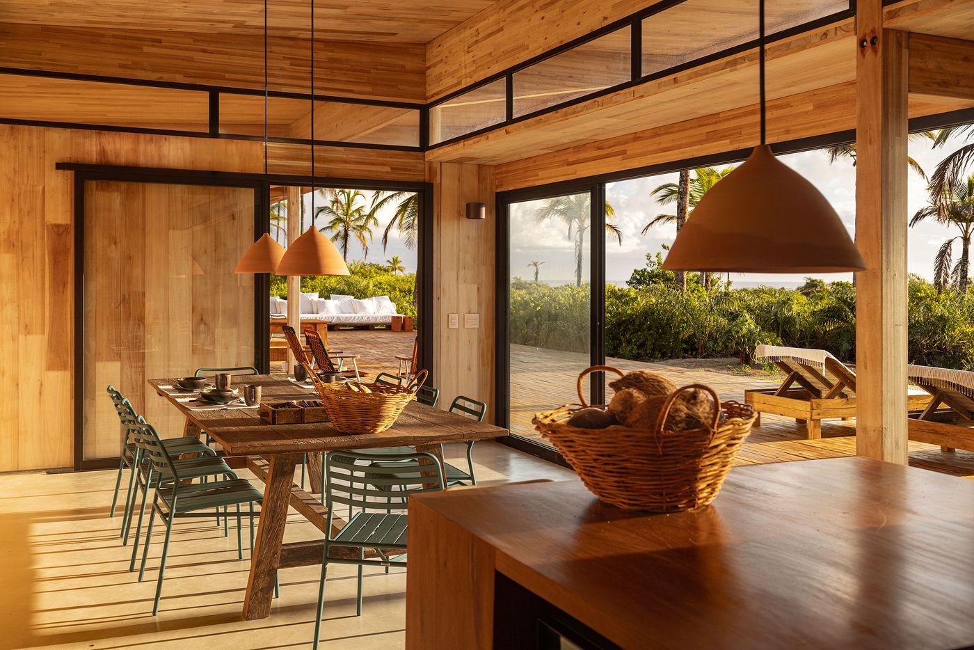 A contemporary dining area, defined by warm wooden ceilings and walls, features a rustic timber table paired with modern green chairs. Large sliding glass doors open to an outdoor deck and tropical landscape, creating a seamless transition and emphasizing a relaxed, indoor-outdoor lifestyle.