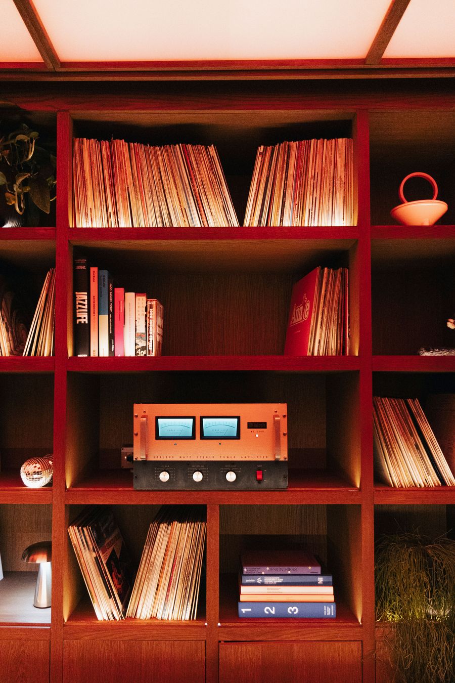 A cozy shelf displaying vinyl records, books, and a vintage audio amplifier with blue meters, illuminated with warm lighting. A cozy shelf displaying vinyl records, books, and a vintage audio amplifier with blue meters, illuminated with warm lighting.