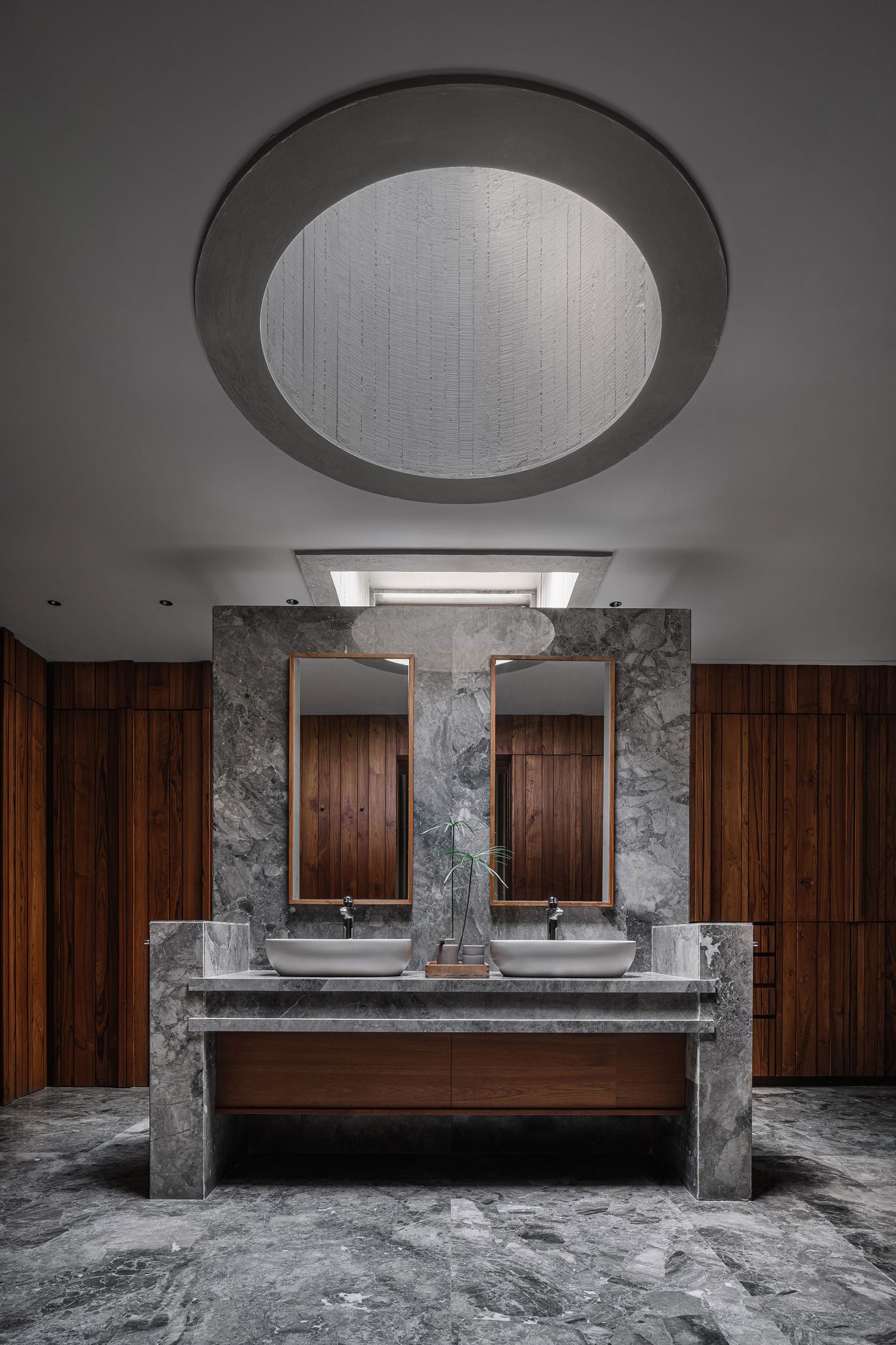 A luxurious and modern bathroom features a double vanity crafted from light grey veined marble, complemented by contemporary vessel sinks. Warm wooden paneled walls provide a striking contrast. An impressive circular skylight in the concrete ceiling bathes the space in natural light, enhancing the sophisticated material palette and minimalist design.
