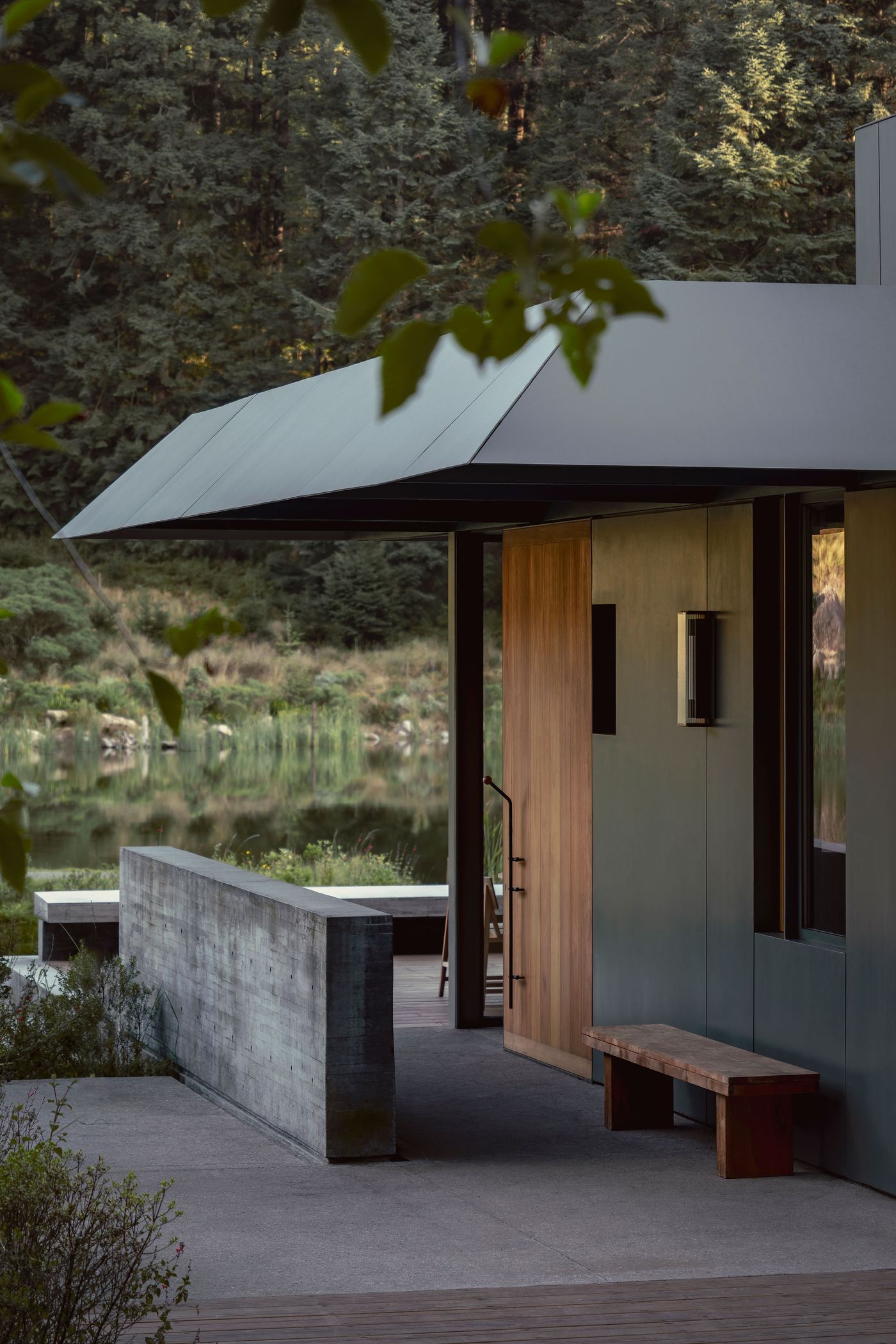 Modern architectural entrance with wooden door, concrete bench, and sleek overhang, surrounded by lush greenery and a tranquil water view.