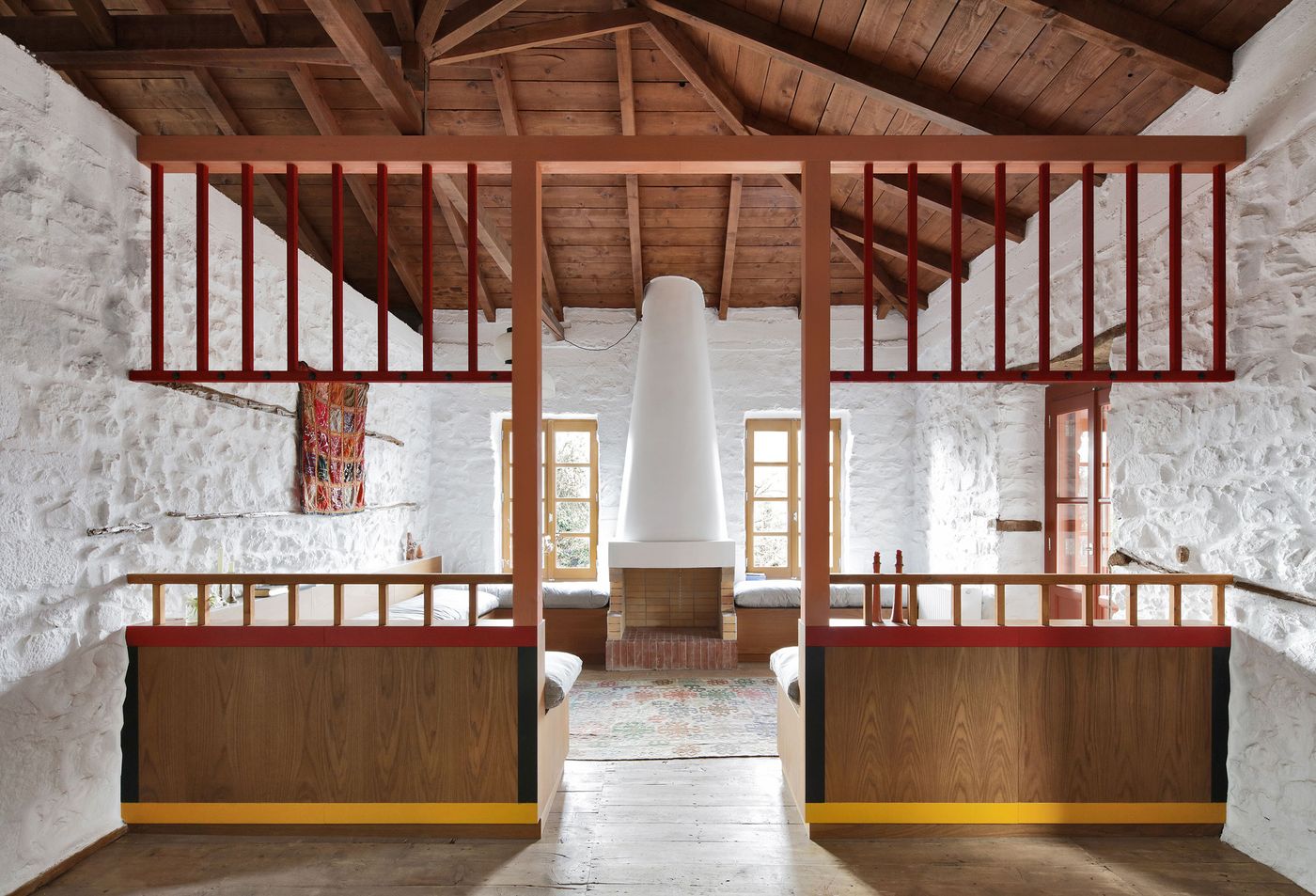 Neo-vernacular restoration creates a communal sleeping hall where coral-red steel mezzanines float above custom oak bed platforms. The exposed timber roof structure and whitewashed stone walls establish rustic authenticity, while contemporary built-in furniture brings modern comfort to traditional spatial arrangements.