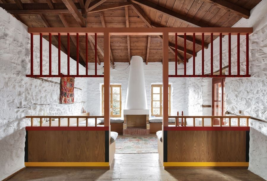 Neo-vernacular restoration creates a communal sleeping hall where coral-red steel mezzanines float above custom oak bed platforms. The exposed timber roof structure and whitewashed stone walls establish rustic authenticity, while contemporary built-in furniture brings modern comfort to traditional spatial arrangements.