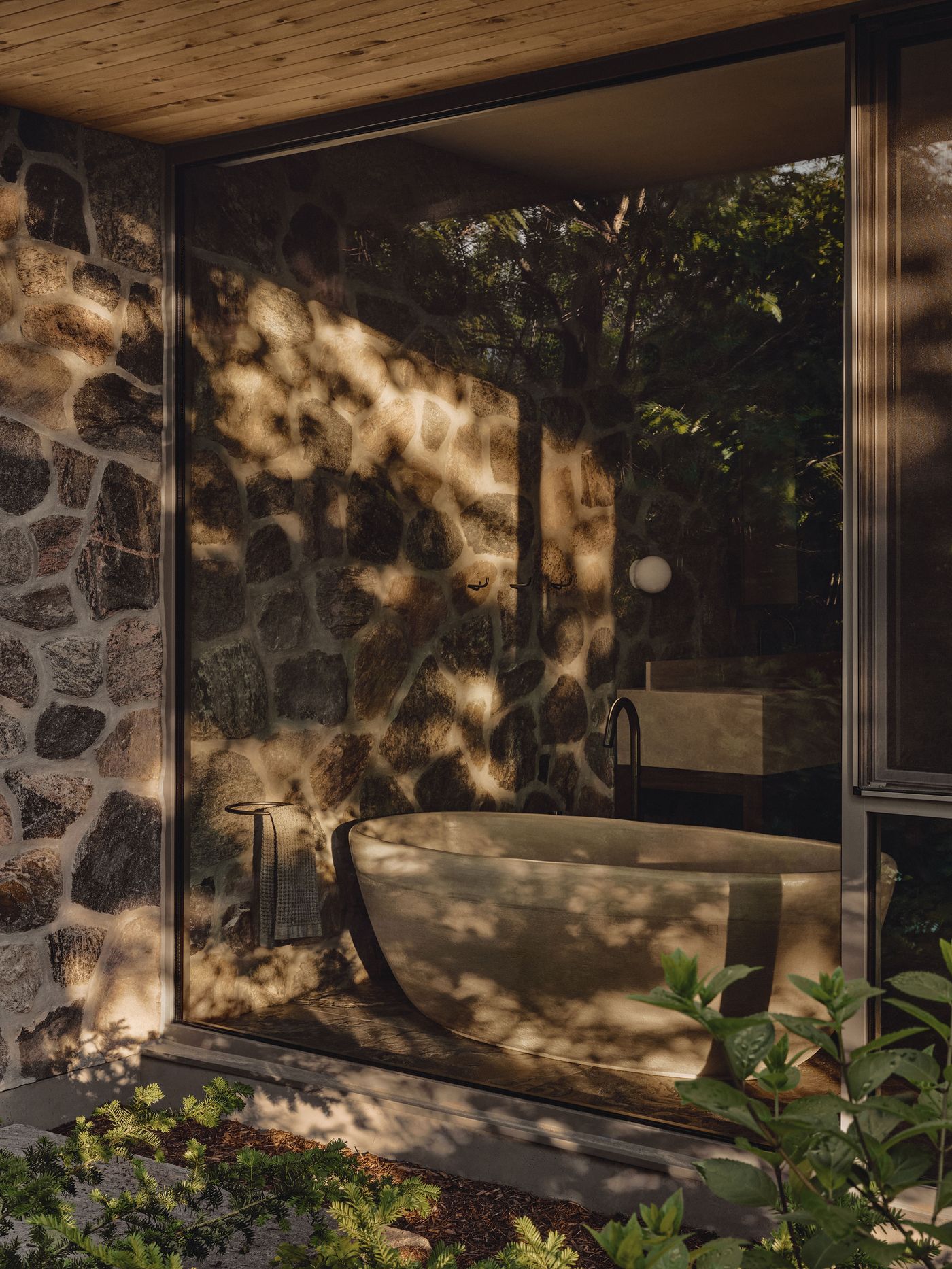 A serene bathroom features a freestanding tub, stone walls, and reflections of greenery, illuminated by warm natural light. A serene bathroom features a freestanding tub, stone walls, and reflections of greenery, illuminated by warm natural light.