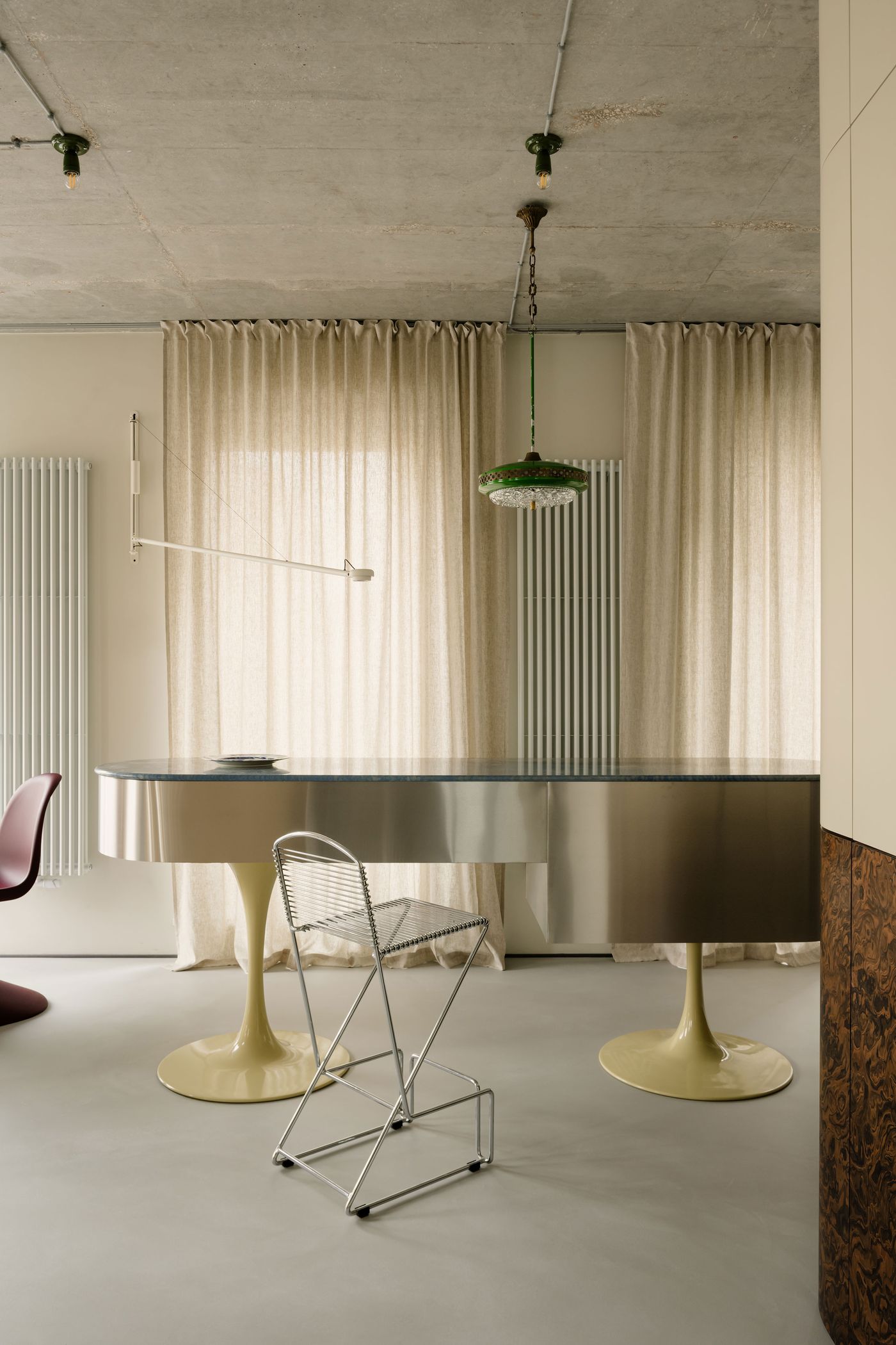 A graceful balance of symmetry and asymmetry is on display in this dining-kitchen hybrid, where a custom piano-shaped island gleams beneath a vintage pendant, flanked by minimalist seating in metal and burgundy plastic.