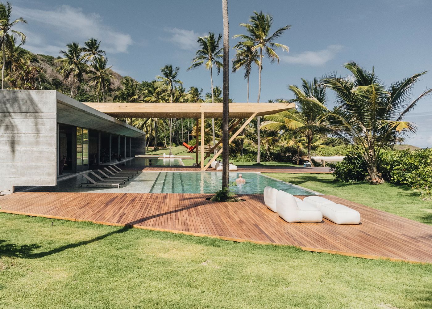 Modern tropical villa with a swimming pool, surrounded by palm trees and a wooden deck, featuring lounge chairs and a person swimming.