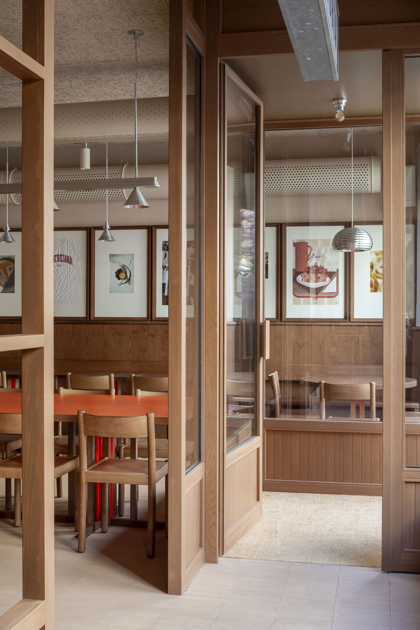 An elegant view through the wooden-framed glass doorways of Santo Palato, designed by Naessi Studio. The composition reveals multiple dining areas, characterized by warm wood paneling, minimalist pendant lights, and framed artwork. The interplay of transparency and solid forms creates a sophisticated yet welcoming atmosphere in this modern Roman trattoria.