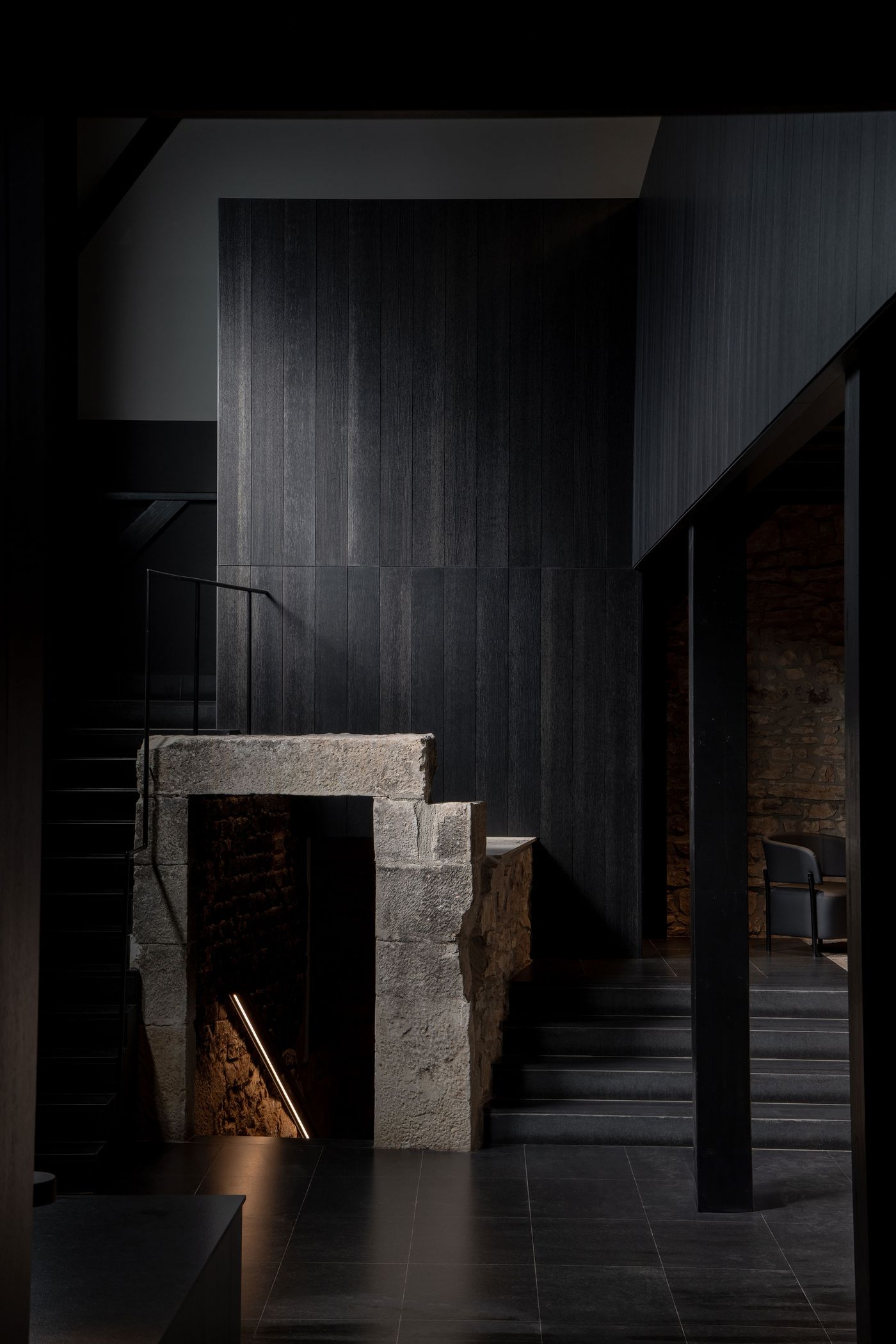 A modern, dark interior features a blend of textured wood and stone elements, with a staircase leading to a softly lit lower level.