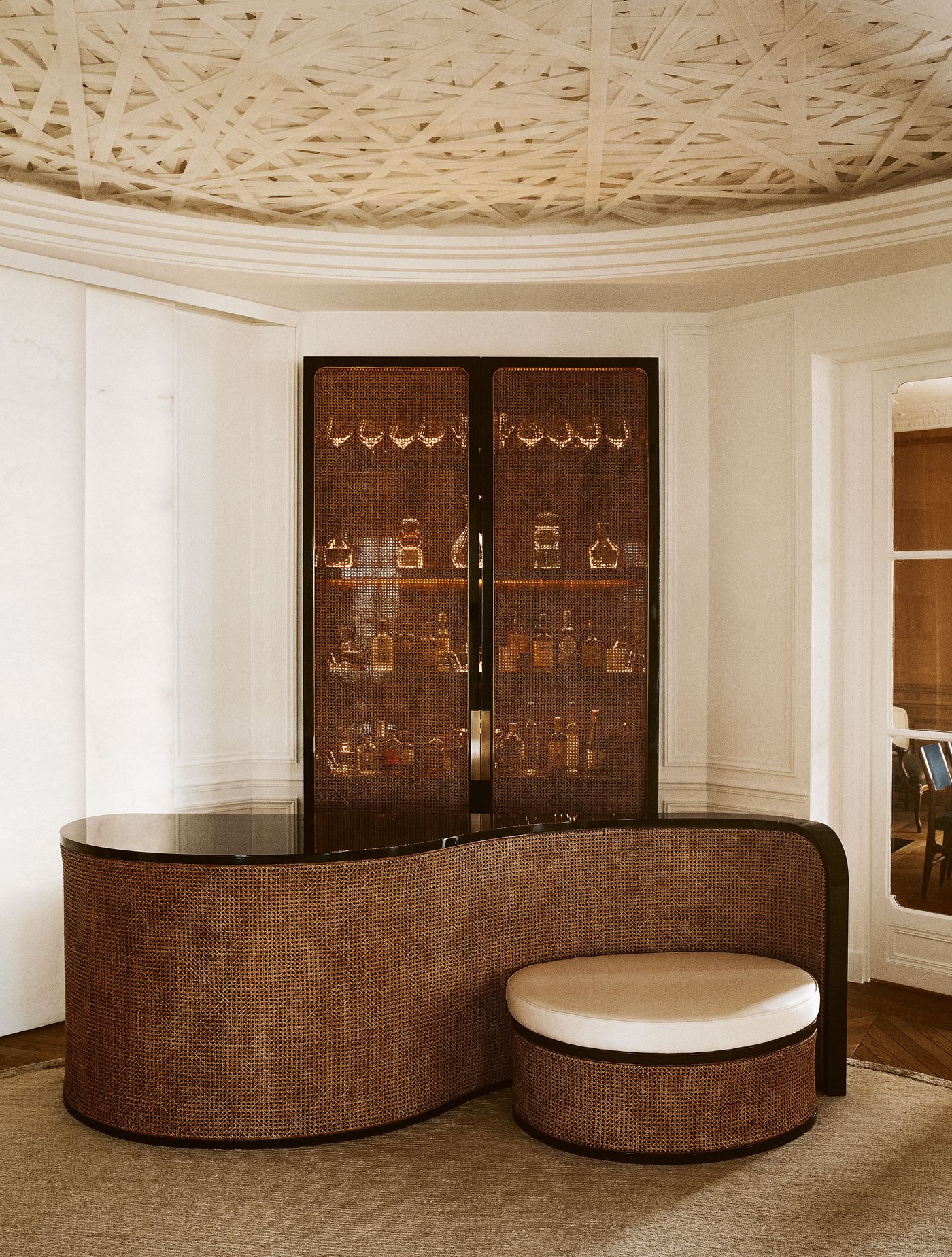 A curved, richly textured rattan bar unit with a dark countertop and a matching cylindrical stool creates a focal point, echoing the artful ceiling texture above. Behind it, a tall, dark display cabinet with woven cane doors reveals an array of glassware, showcasing a fusion of natural materials and elegant design.