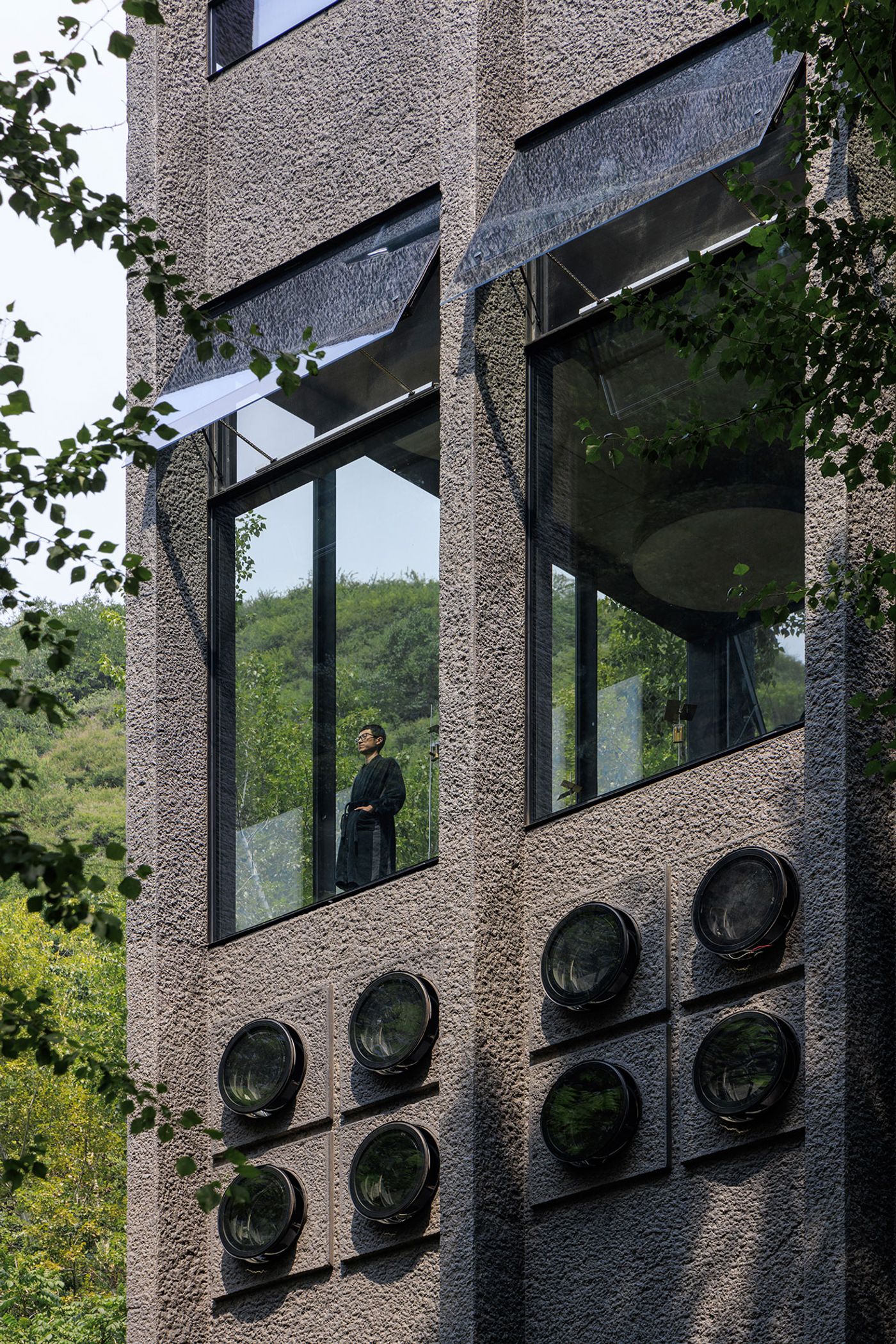 A modern, concrete-textured building with large glass windows, featuring round accents on the exterior and surrounded by greenery. A modern, concrete-textured building with large glass windows, featuring round accents on the exterior and surrounded by greenery.