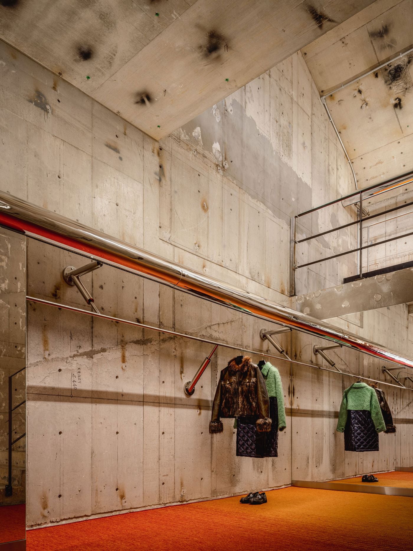 Clothing hangs directly against the exposed concrete shell, suspended from slender chrome rails beneath a polished metal tube that stretches along the wall. Orange-red flooring adds warmth at ground level, while mirrored panels duplicate the garments and architecture, giving the compact vignette greater depth and visual ambiguity.