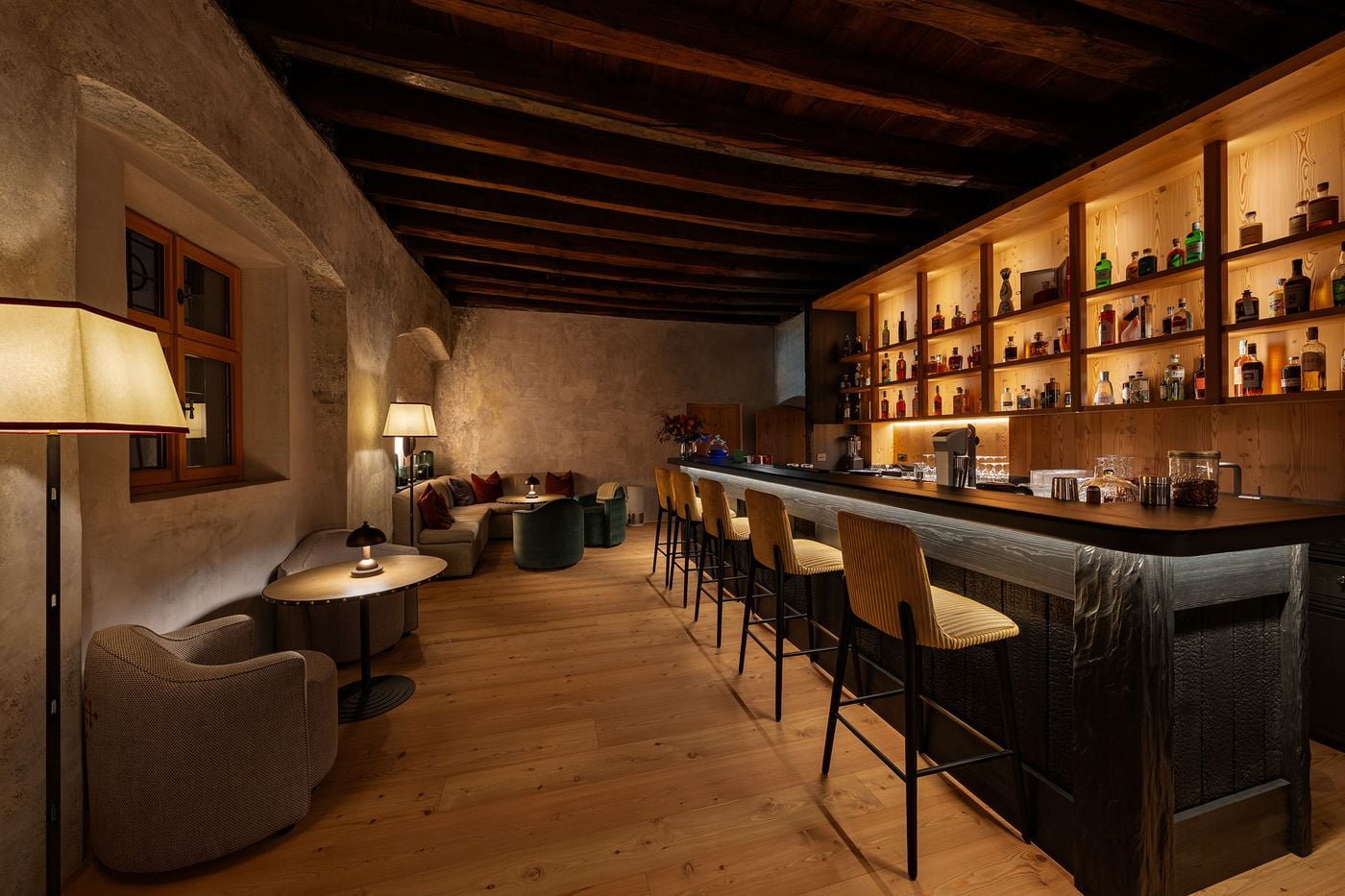 Castel Badia's bar sets backlit timber shelving and a counter clad in charred, deeply textured wood beneath a dark beamed ceiling. Yellow striped bar stools line the counter, while mottled plaster walls, pleated lampshades, green velvet tub chairs and a deep banquette shape a convivial lounge beyond. Castel Badia's bar sets backlit timber shelving and a counter clad in charred, deeply textured wood beneath a dark beamed ceiling. Yellow striped bar stools line the counter, while mottled plaster walls, pleated lampshades, green velvet tub chairs and a deep banquette shape a convivial lounge beyond.