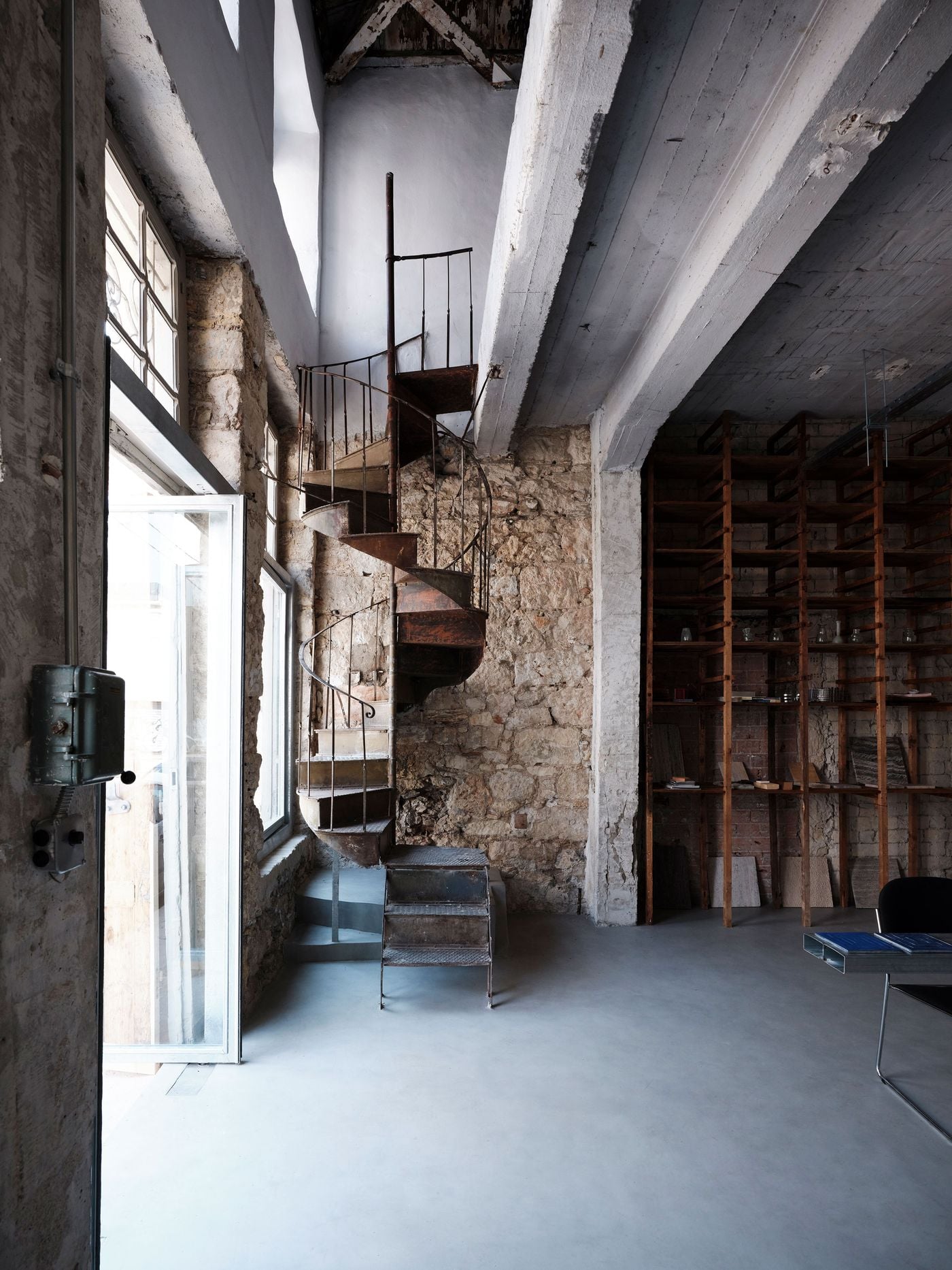 The original rusted spiral staircase rises dramatically against rough stonework, accompanied by a small metal stair bridging its first step. Nearby, a tall vintage timber storage unit has been repurposed for books and material samples, retaining another utilitarian remnant of the building’s former workshop life. The original rusted spiral staircase rises dramatically against rough stonework, accompanied by a small metal stair bridging its first step. Nearby, a tall vintage timber storage unit has been repurposed for books and material samples, retaining another utilitarian remnant of the building’s former workshop life.