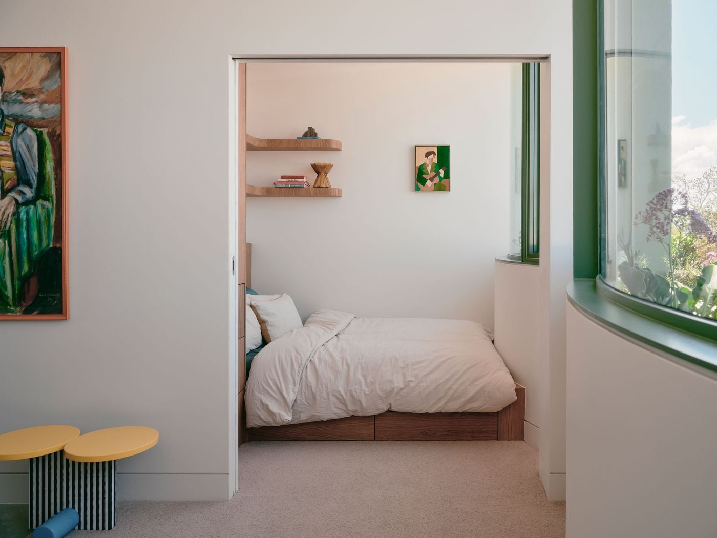 A wide opening connects the living area to a compact bedroom, where a custom timber bed with integrated storage sits beneath floating shelves and a small artwork. The curved sage-green window bay draws daylight deep into the room, while playful yellow side tables in the foreground echo the project’s colourful exterior accents. A wide opening connects the living area to a compact bedroom, where a custom timber bed with integrated storage sits beneath floating shelves and a small artwork. The curved sage-green window bay draws daylight deep into the room, while playful yellow side tables in the foreground echo the project’s colourful exterior accents.