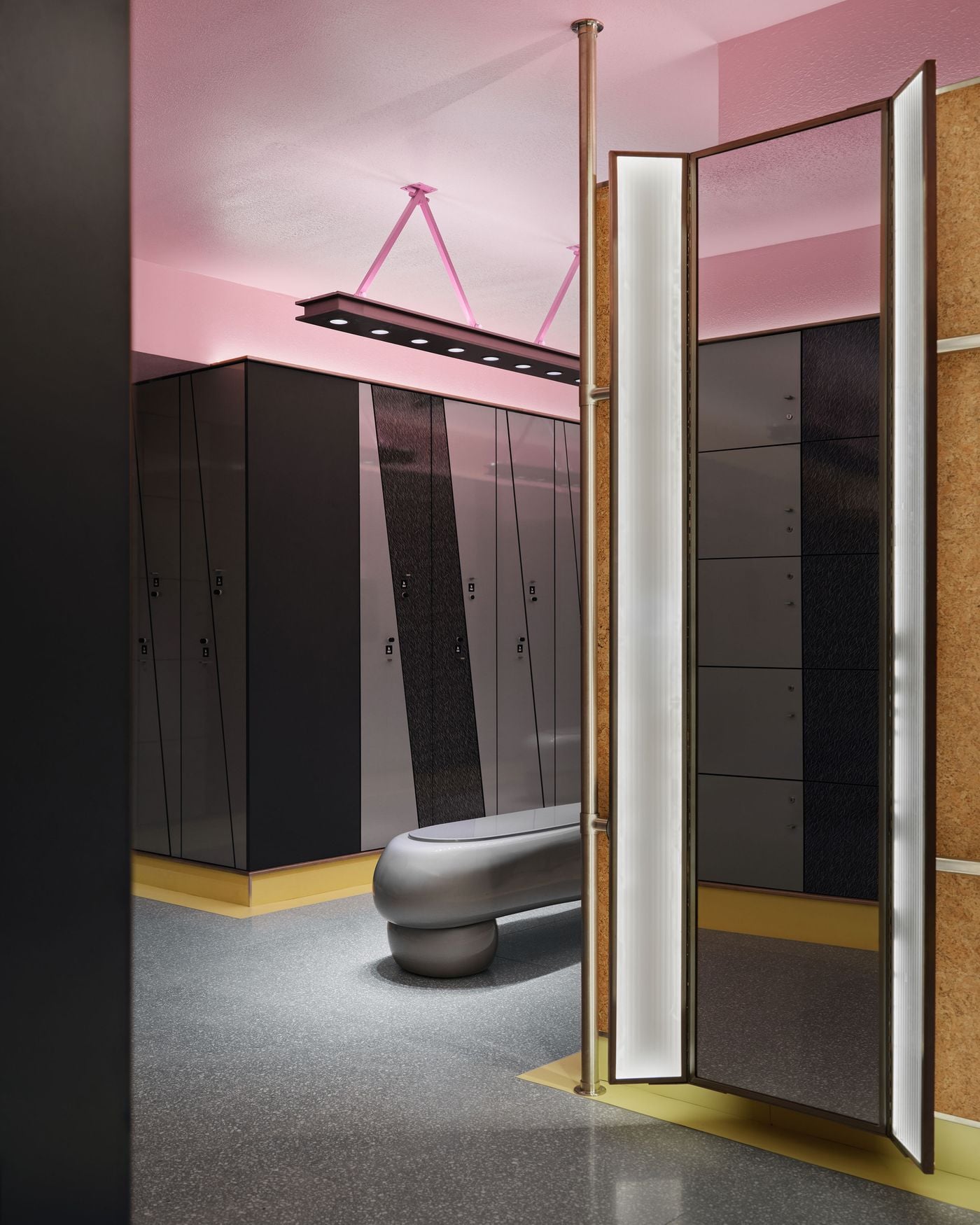 The men’s locker room reverses conventional colour coding with a pink ceiling, while dark metal lockers, cork-lined walls and a sculptural silver bench create a theatrical setting. A freestanding illuminated mirror screen adds another graphic layer, framed by stainless-steel poles and yellow skirting. The men’s locker room reverses conventional colour coding with a pink ceiling, while dark metal lockers, cork-lined walls and a sculptural silver bench create a theatrical setting. A freestanding illuminated mirror screen adds another graphic layer, framed by stainless-steel poles and yellow skirting.