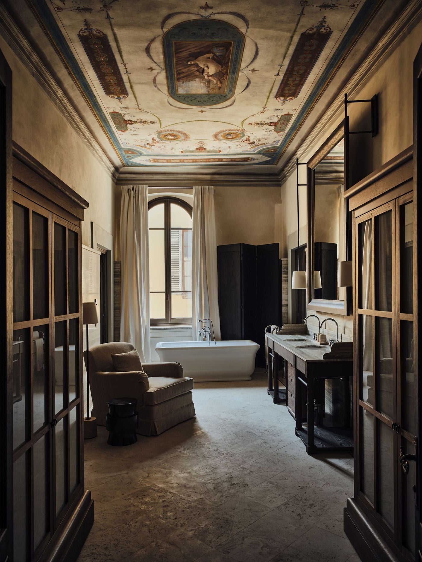A painted ceiling with a central mythological scene presides over a long bathroom at La Réserve Firenze. Glazed oak cabinets frame the view towards a freestanding bathtub beneath an arched window, accompanied by a skirted armchair, a dark folding screen and a double vanity in travertine and oak. A painted ceiling with a central mythological scene presides over a long bathroom at La Réserve Firenze. Glazed oak cabinets frame the view towards a freestanding bathtub beneath an arched window, accompanied by a skirted armchair, a dark folding screen and a double vanity in travertine and oak.