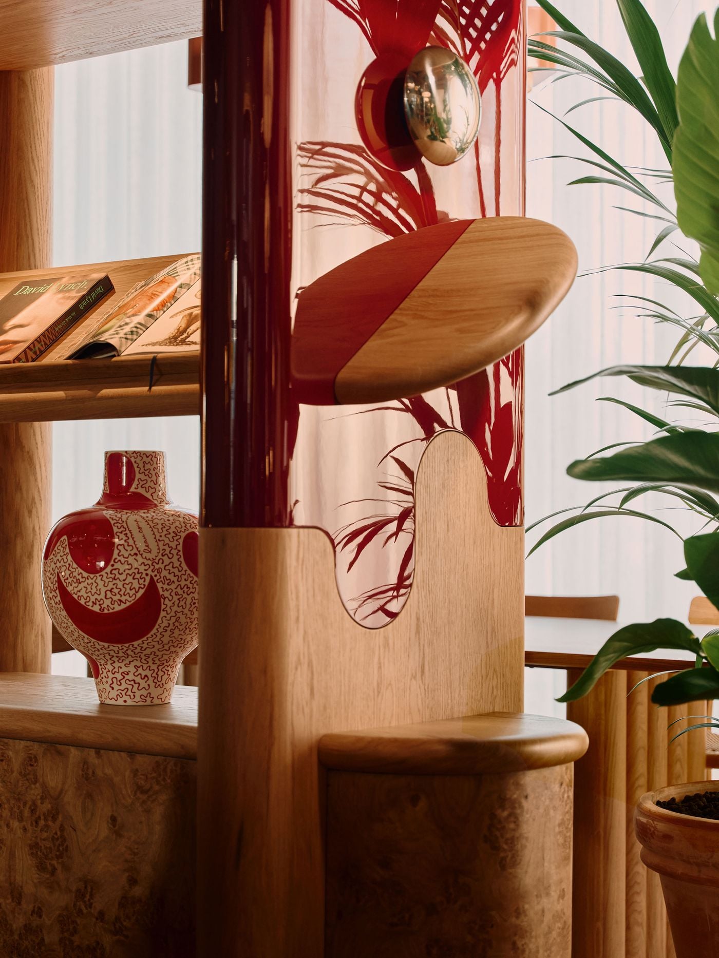 A close-up reveals the crafted detailing of the bespoke lounge joinery, where a glossy burgundy panel meets pale oak in an oval pivoting shelf. Reflections of surrounding foliage animate the lacquered surface, while a patterned ceramic vase and nearby planting heighten the tactile, almost domestic quality of Holloway Li’s design. A close-up reveals the crafted detailing of the bespoke lounge joinery, where a glossy burgundy panel meets pale oak in an oval pivoting shelf. Reflections of surrounding foliage animate the lacquered surface, while a patterned ceramic vase and nearby planting heighten the tactile, almost domestic quality of Holloway Li’s design.