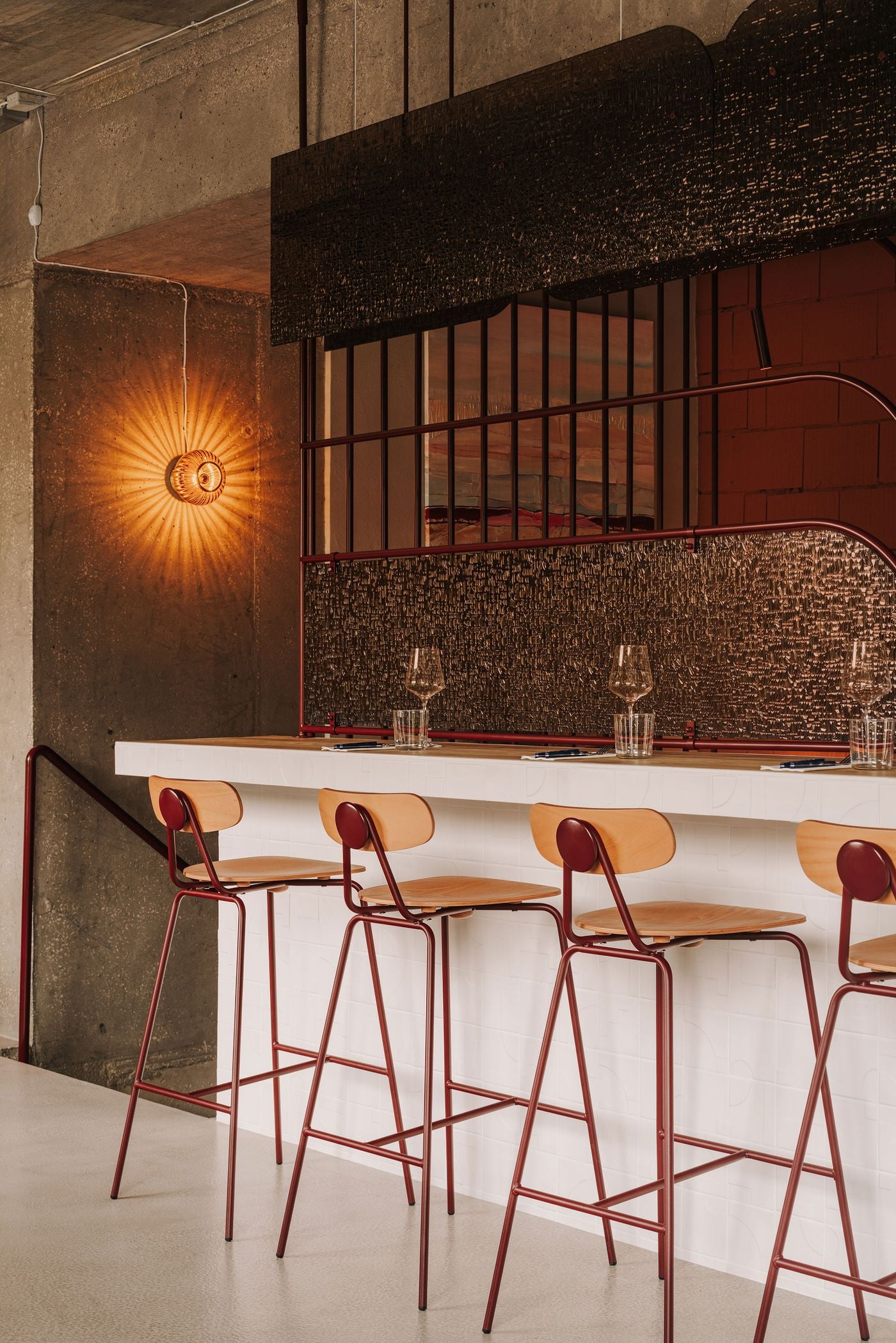 A closer perspective of WIN’s upstairs dining counter highlights the interplay of white relief-patterned ceramic tiles, warm timber and burgundy-framed bentwood stools. Behind the counter, slender vertical rods and undulating metal rails frame textured glass panels, while a ribbed wall sconce casts radiating light across exposed concrete, accentuating the contrast between industrial materials and decorative detailing. A closer perspective of WIN’s upstairs dining counter highlights the interplay of white relief-patterned ceramic tiles, warm timber and burgundy-framed bentwood stools. Behind the counter, slender vertical rods and undulating metal rails frame textured glass panels, while a ribbed wall sconce casts radiating light across exposed concrete, accentuating the contrast between industrial materials and decorative detailing.