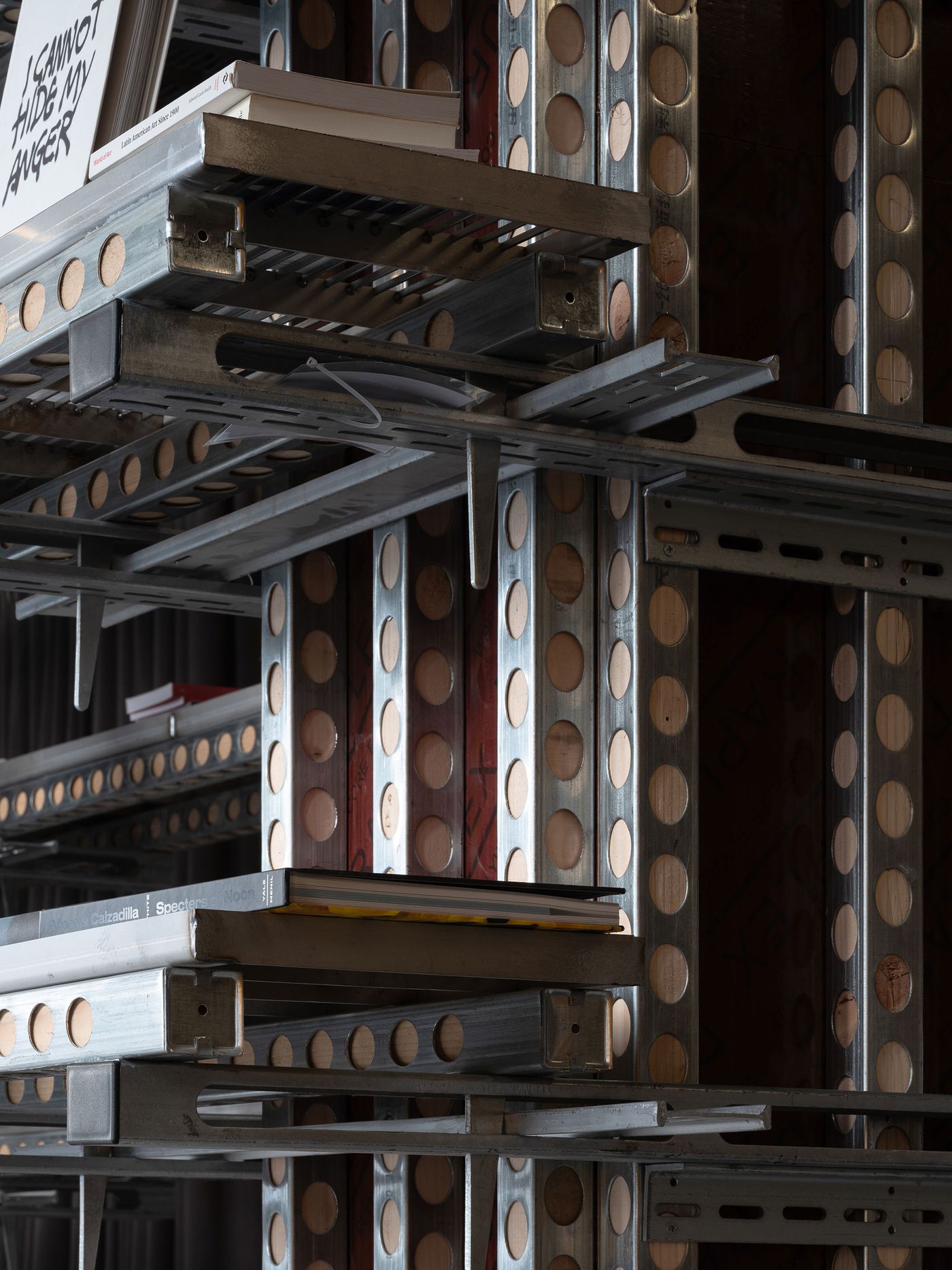 A close-up of the industrial shelving in 889 GLO Art Space emphasizes its intricate, perforated metal structure and the robust, dark-paneled uprights. Various art books, including one titled "I CANNOT HIDE MY ANGER," are neatly arranged, showcasing how the raw, modular design serves as a sophisticated display system within this brutalist-inspired space.