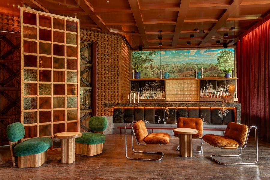 This panoramic view of BEKEB, designed by maye estudio, reveals a sophisticated lounge area. A long bar with a cowhide-covered counter is backed by shelves stocked with bottles and illuminated by warm light. Seating includes plush green armchairs and burnt orange chairs, set against a backdrop of textured walls and dramatic red curtains, all under a richly paneled wooden ceiling.